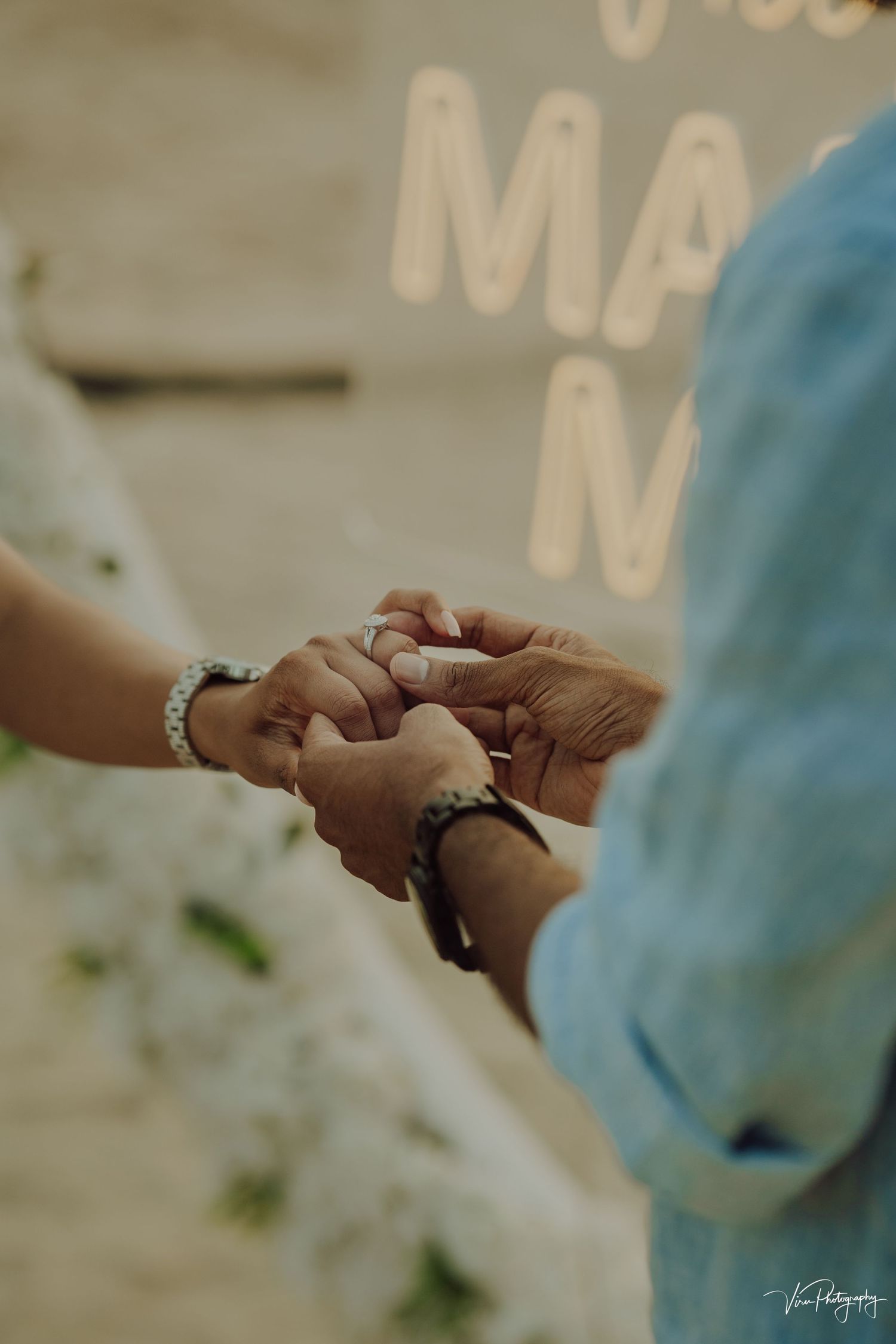 Close-up shot of two people holding hands tenderly with a blurred background at a warm-toned event setting.