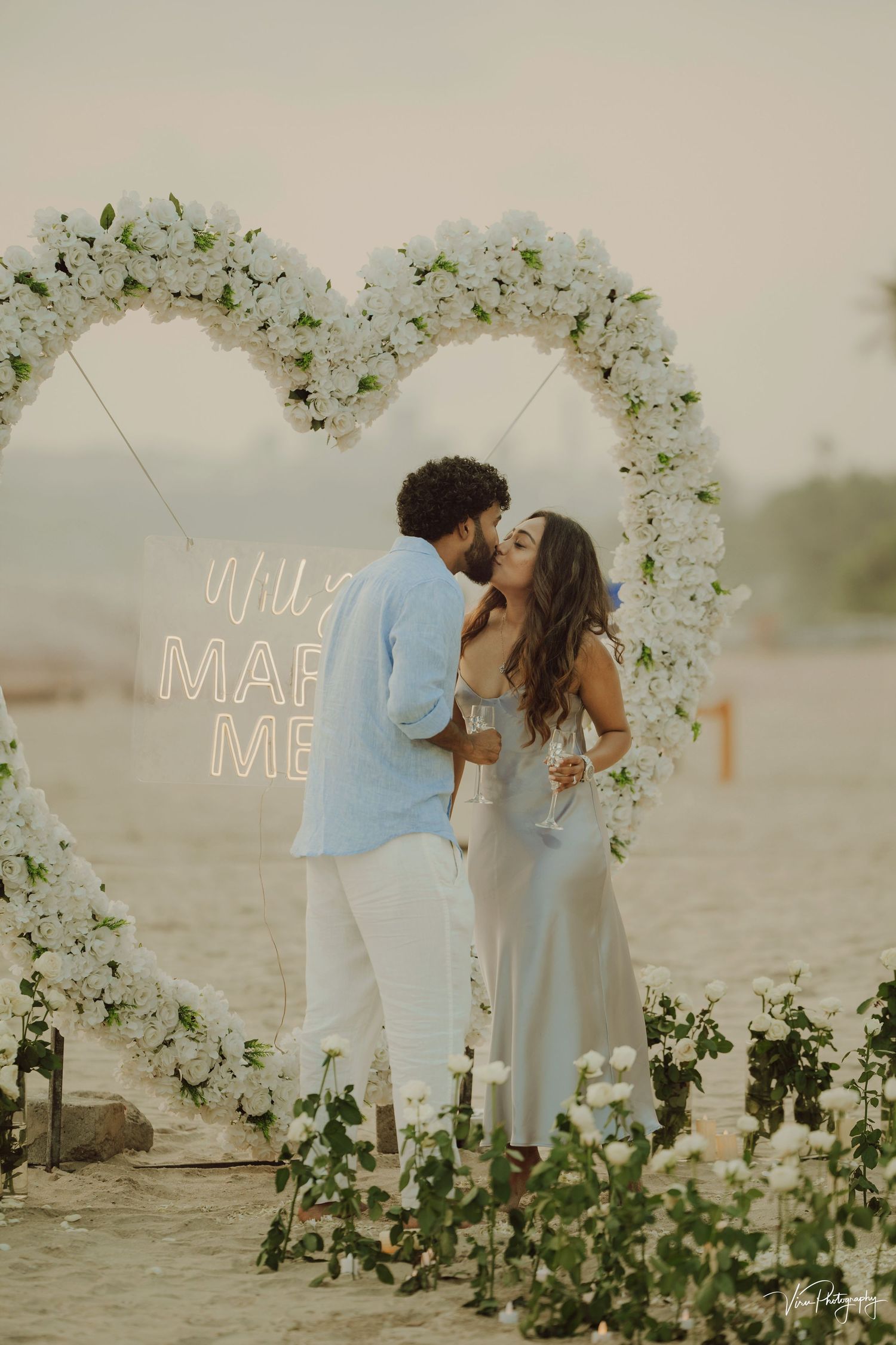A romantic marriage proposal setup with a white floral arch and illuminated 'Marry Me' sign against a natural outdoor backdrop.
