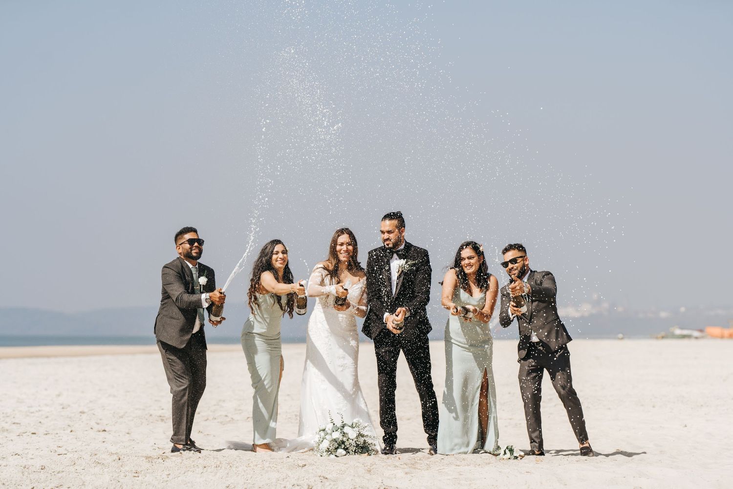 Wedding party wearing gray suits and light dresses poses together on a bright beach with ocean in background.