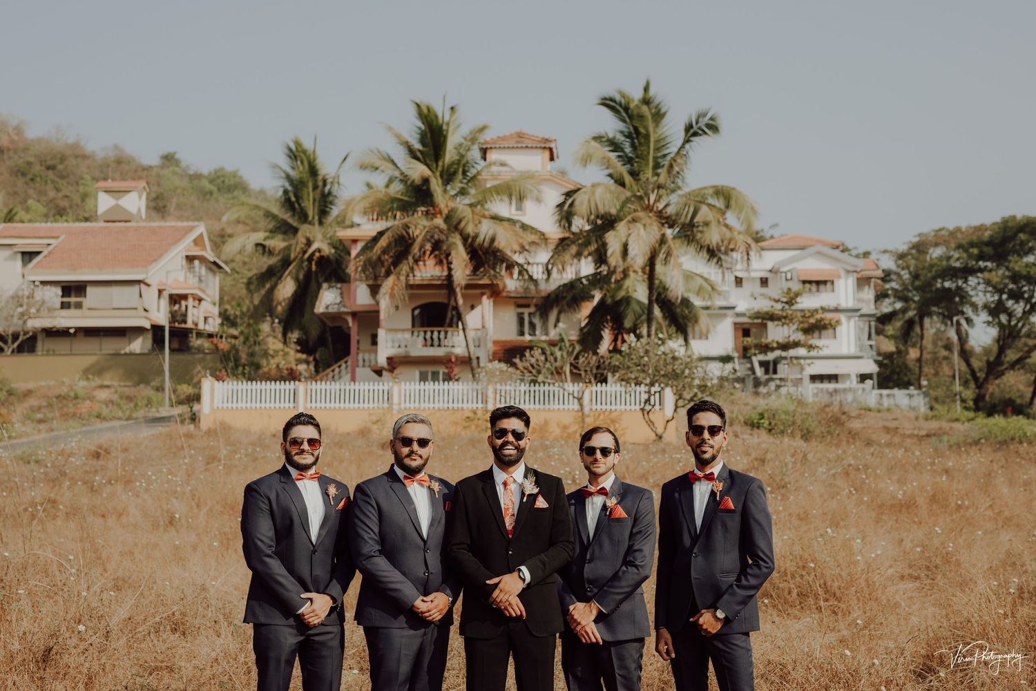 A group in formal suits stands together outdoors with palm trees and houses visible in the background.