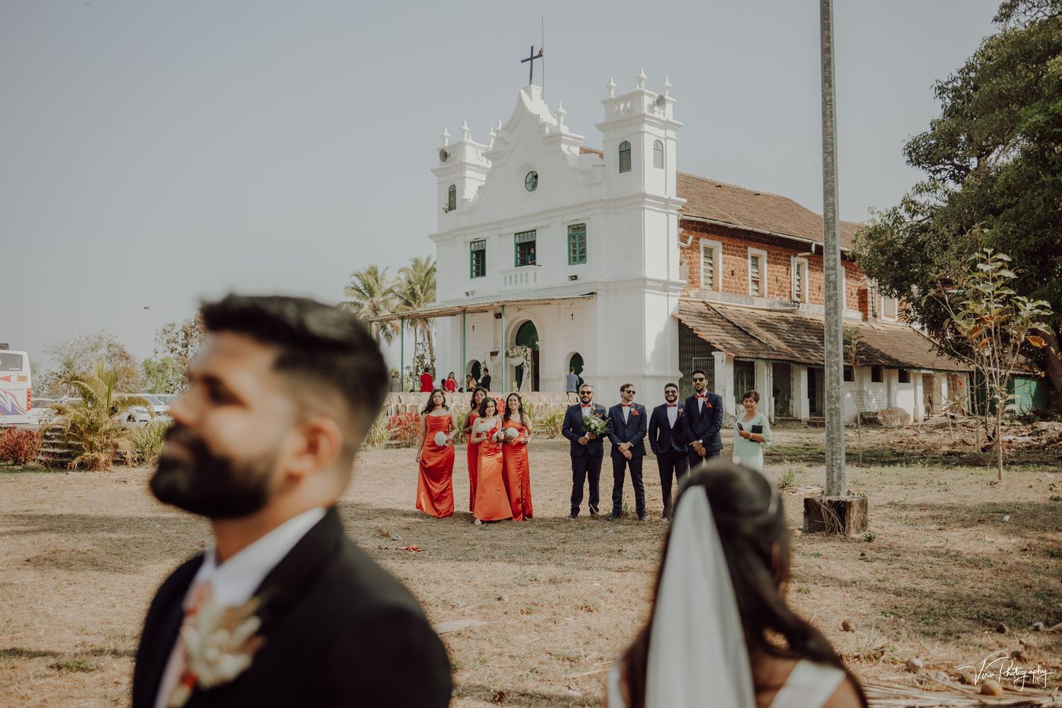 A wedding party stands outside a white colonial-style church with ornate architecture and a cross on top.