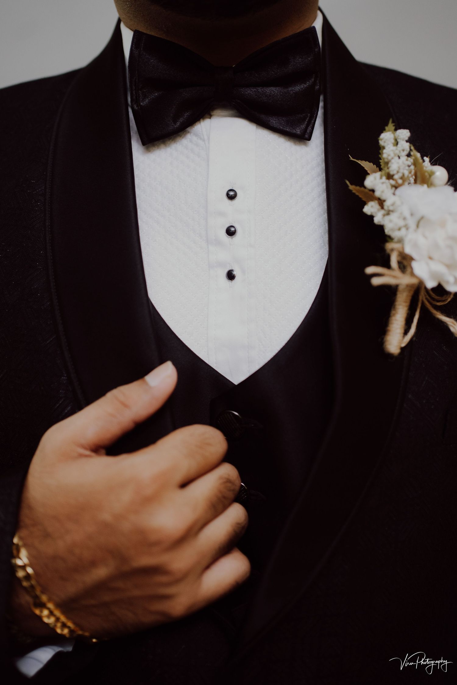 Close-up of formal black tuxedo with white boutonniere, bow tie, and gold watch showing classic wedding attire details.