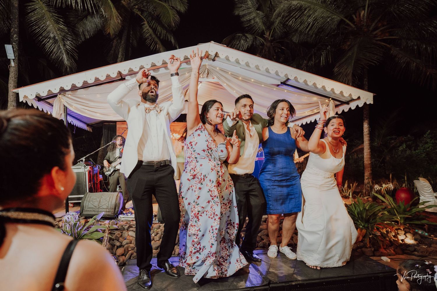A group of celebrants dance together on a covered platform during an outdoor evening wedding reception.