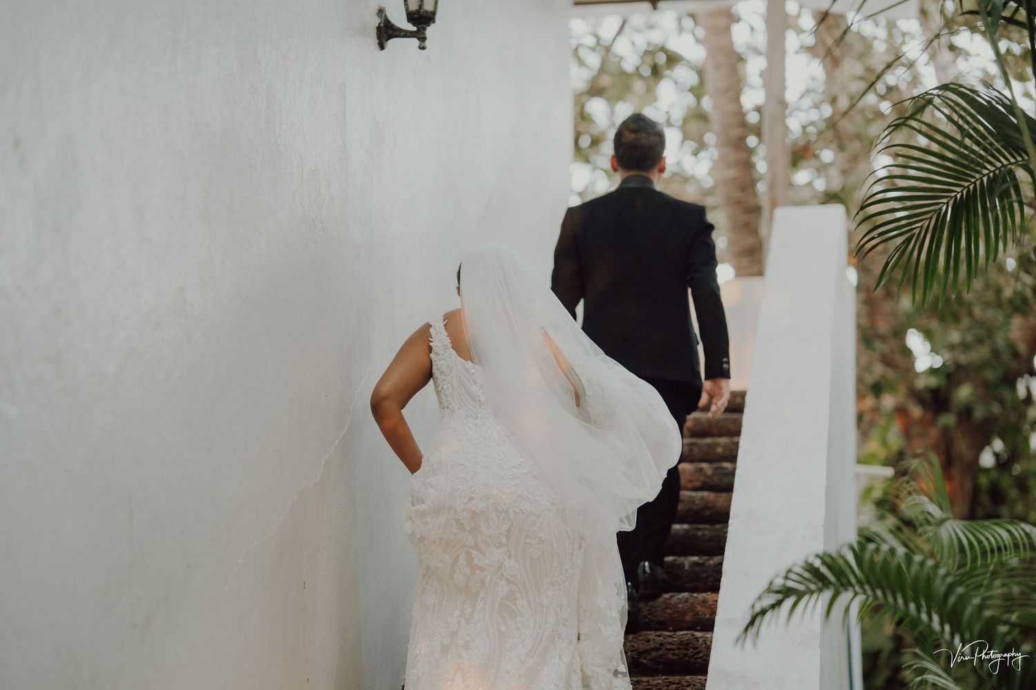 Bride in flowing white gown follows groom up stone steps surrounded by tropical foliage and white walls.