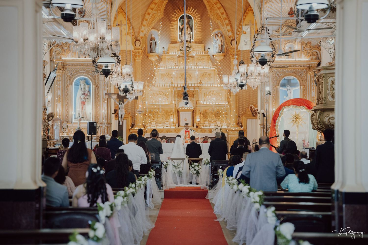 Traditional church wedding ceremony with ornate gold altar, stained glass windows, and white floral aisle decorations.