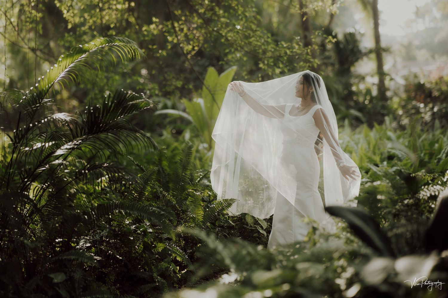 A bride in a flowing white veil poses in a lush tropical garden setting with sunlight filtering through the foliage.