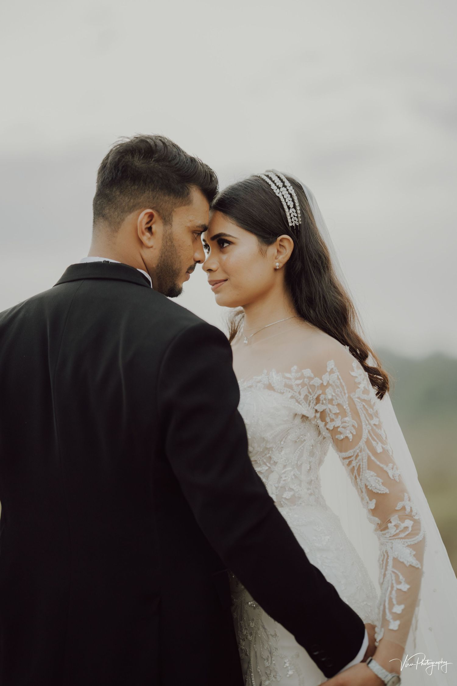 A romantic wedding portrait showing a couple in a black suit and lace wedding dress sharing an intimate moment outdoors.