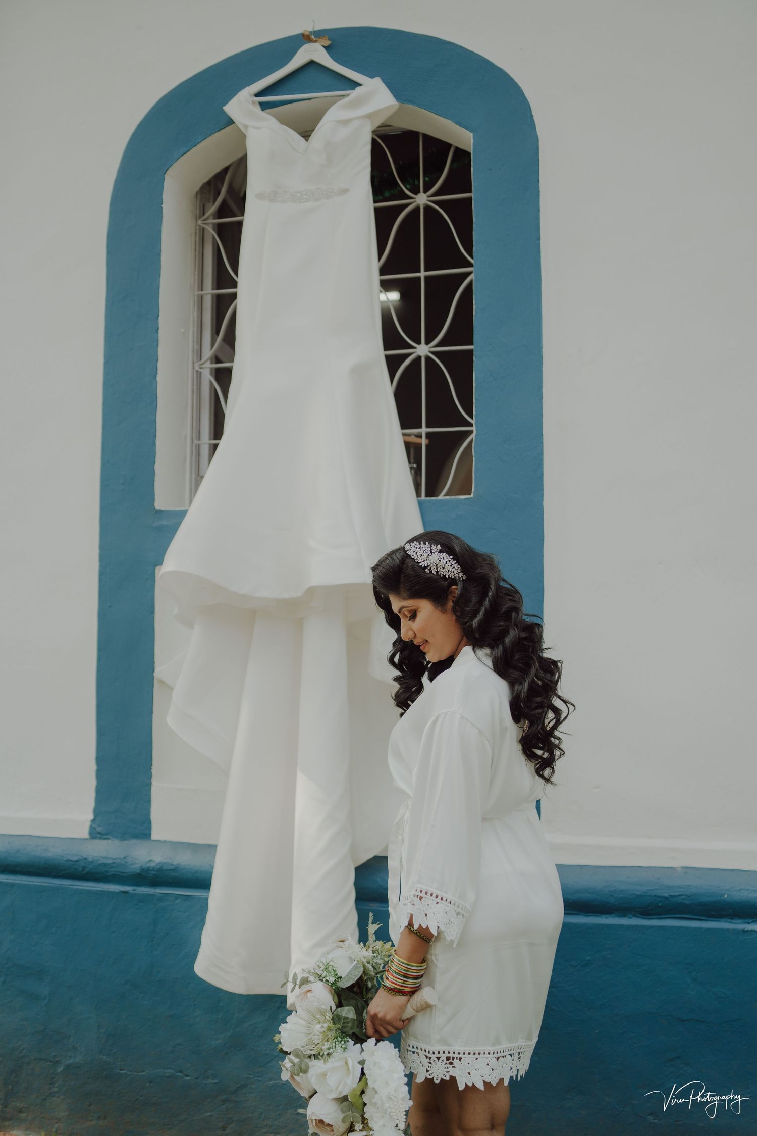 Elegant white wedding dress hangs by arched window while bride in white robe holds bouquet of white flowers.
