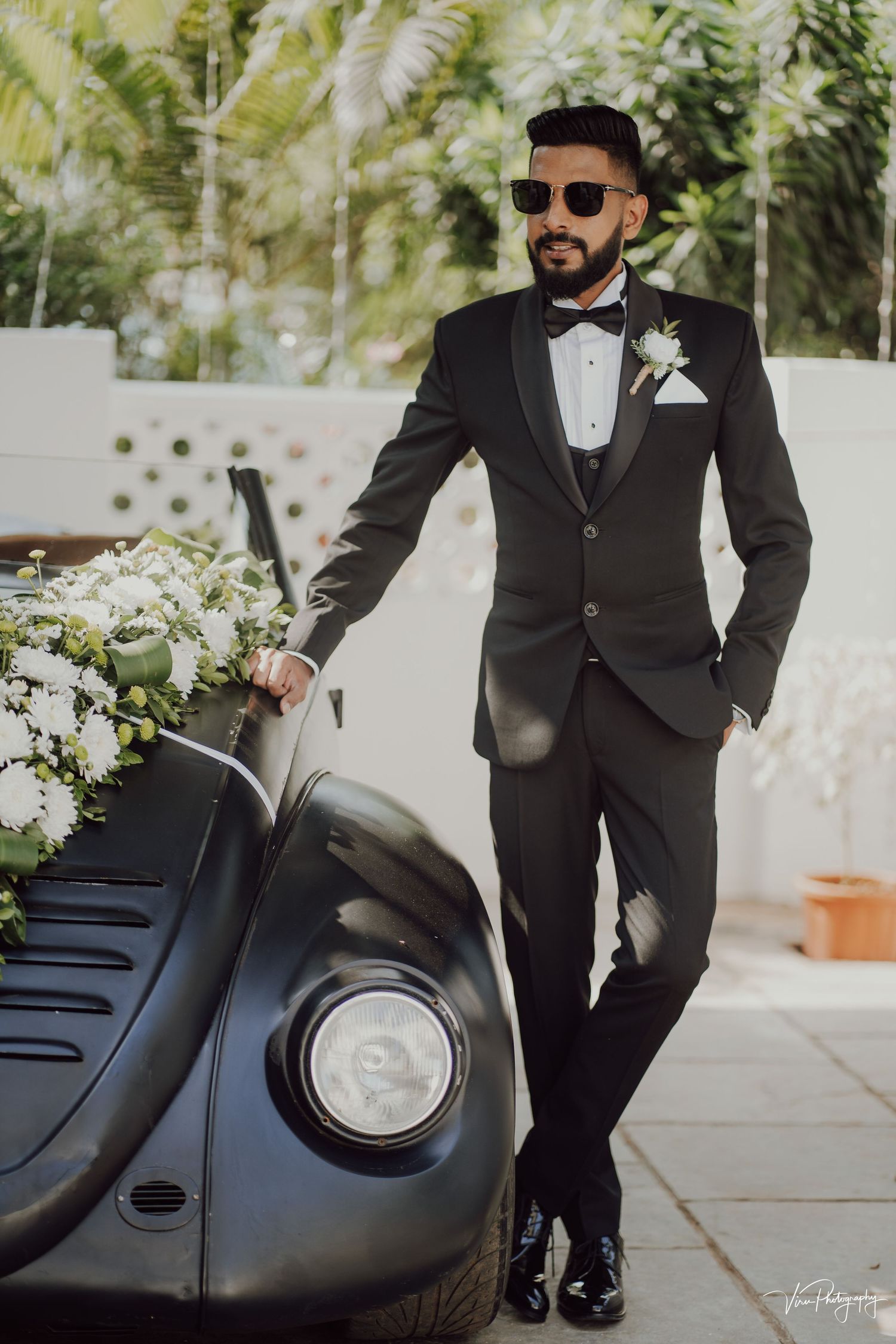 Stylish groom in black tuxedo and sunglasses poses confidently next to vintage black car on wedding day.