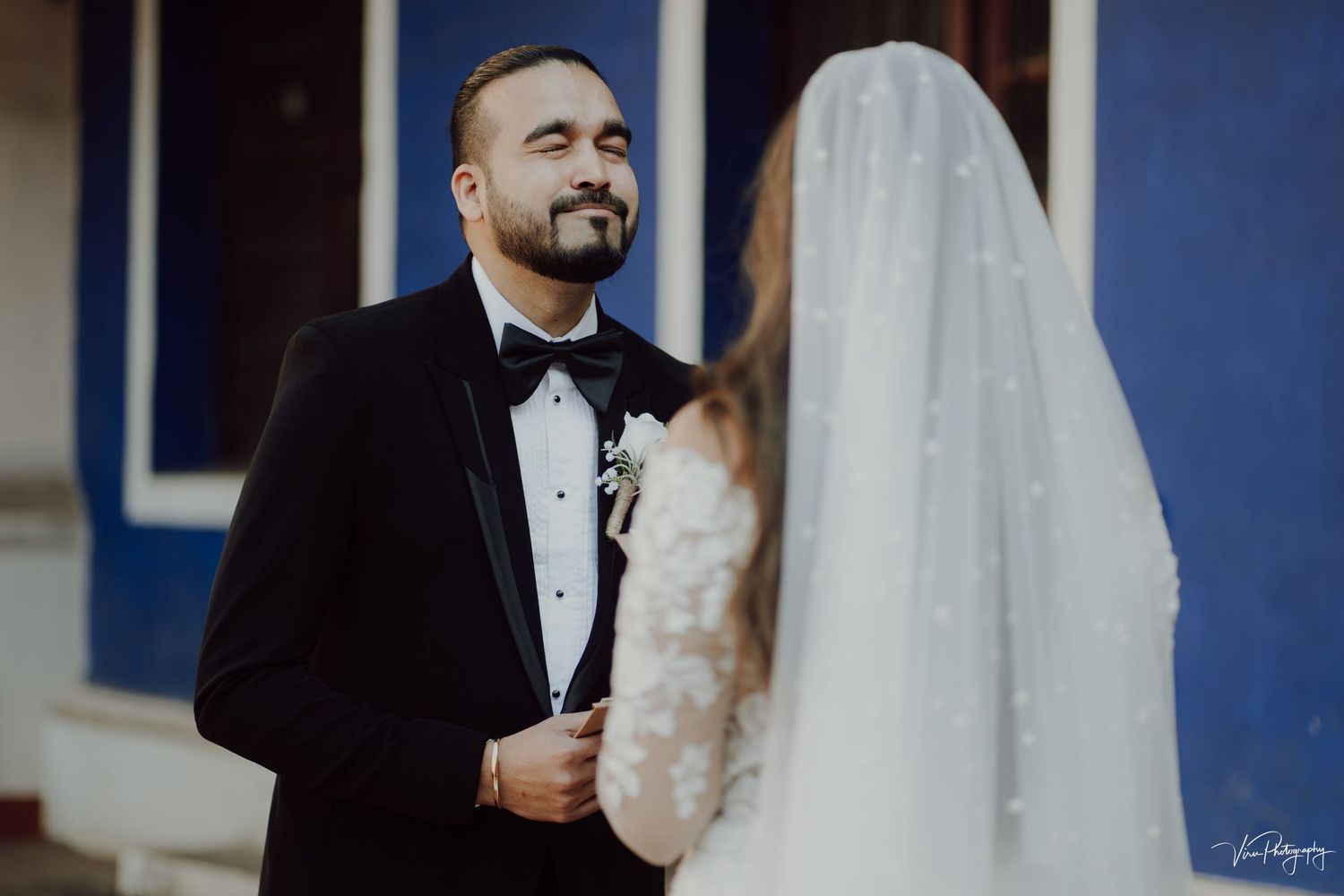 A groom in a black tuxedo and bowtie smiles while standing next to a bride in a white veil at their wedding ceremony.