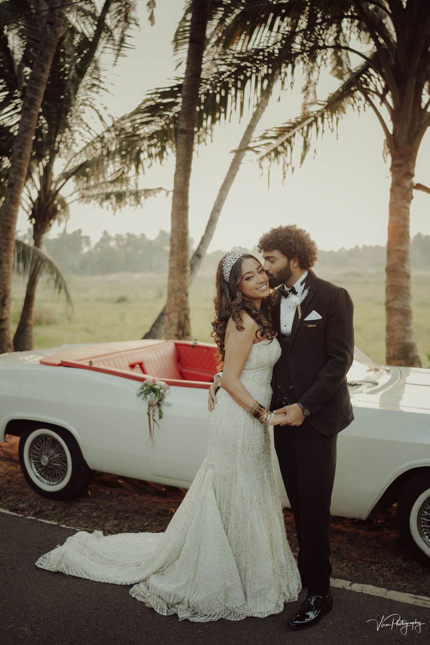 A romantic wedding photo featuring a couple embracing beside a vintage white car under tropical palm trees.