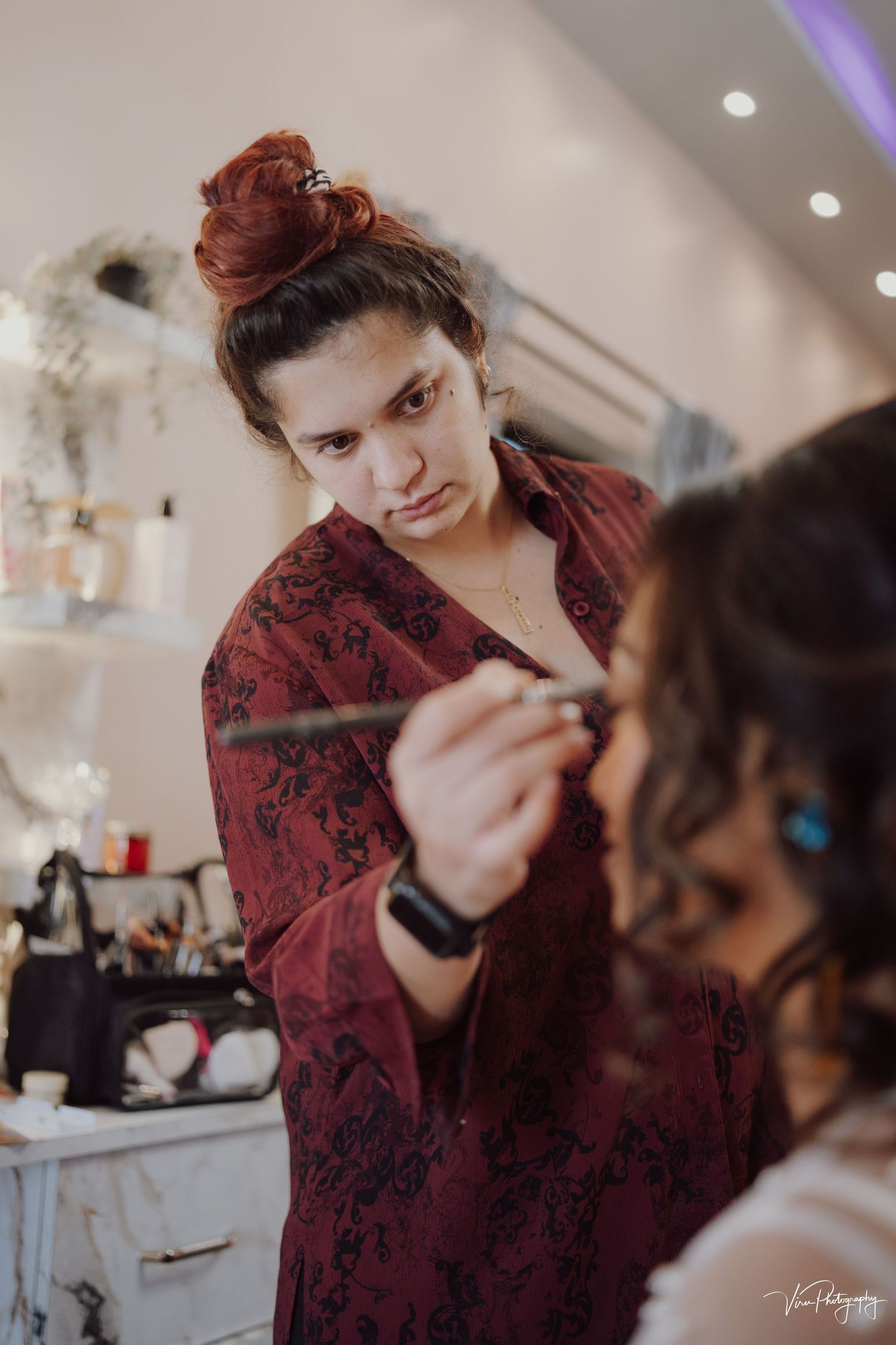 A makeup artist wearing a burgundy floral top applies cosmetics to a client in a beauty salon setting.