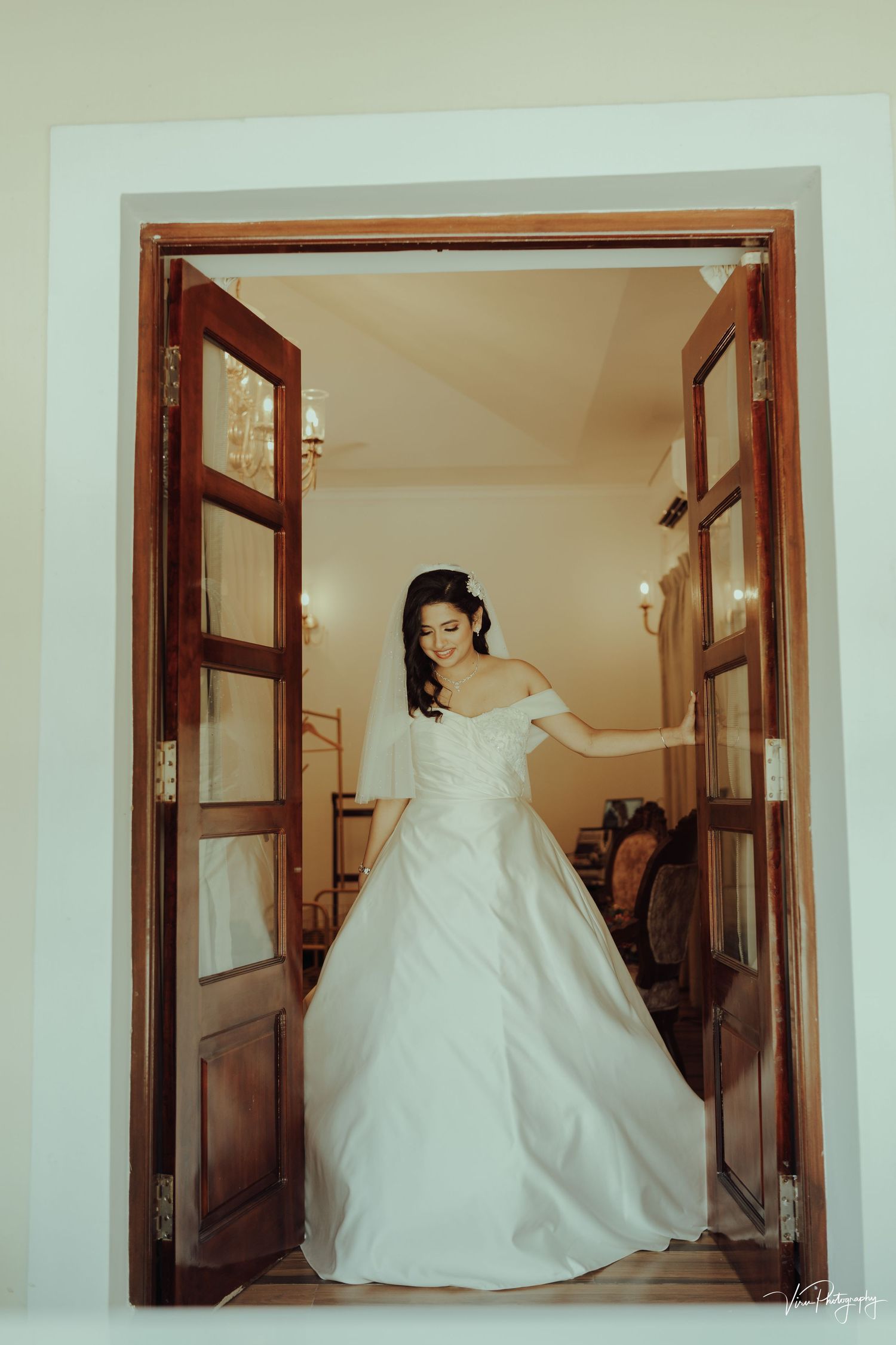 A wedding dress-clad figure stands elegantly framed by wooden double doors in a classic bridal photography pose.