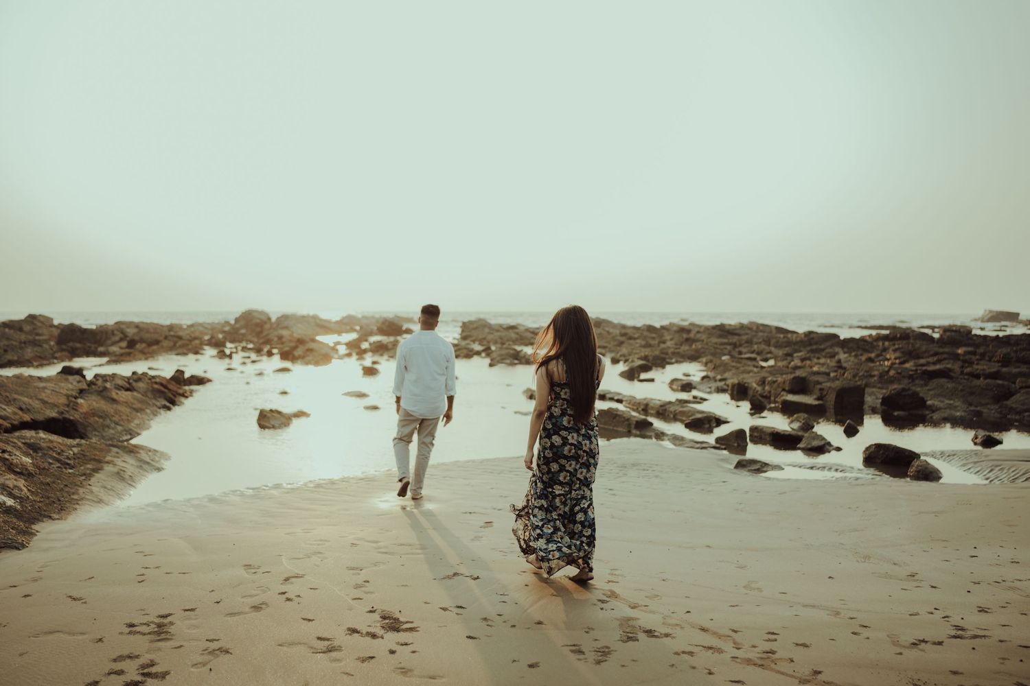 Couple walking on rocky beach shore in vintage-style engagement photo.