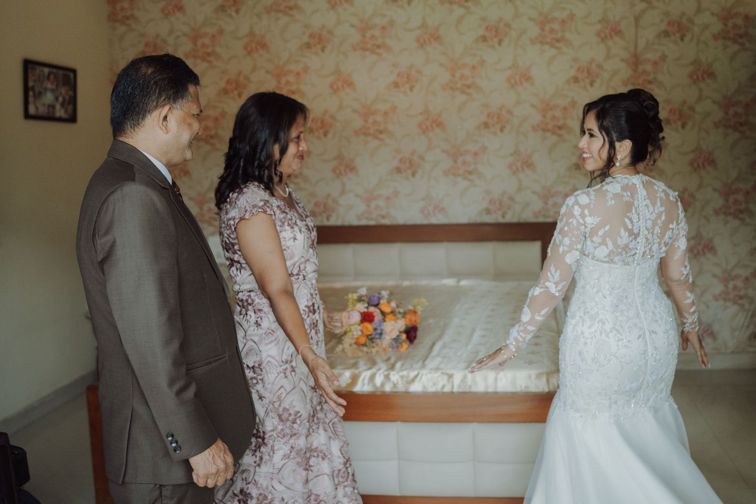 A bride in a lace wedding gown stands before her parents in a bedroom as they look toward her, with a floral bouquet resting on the bed.