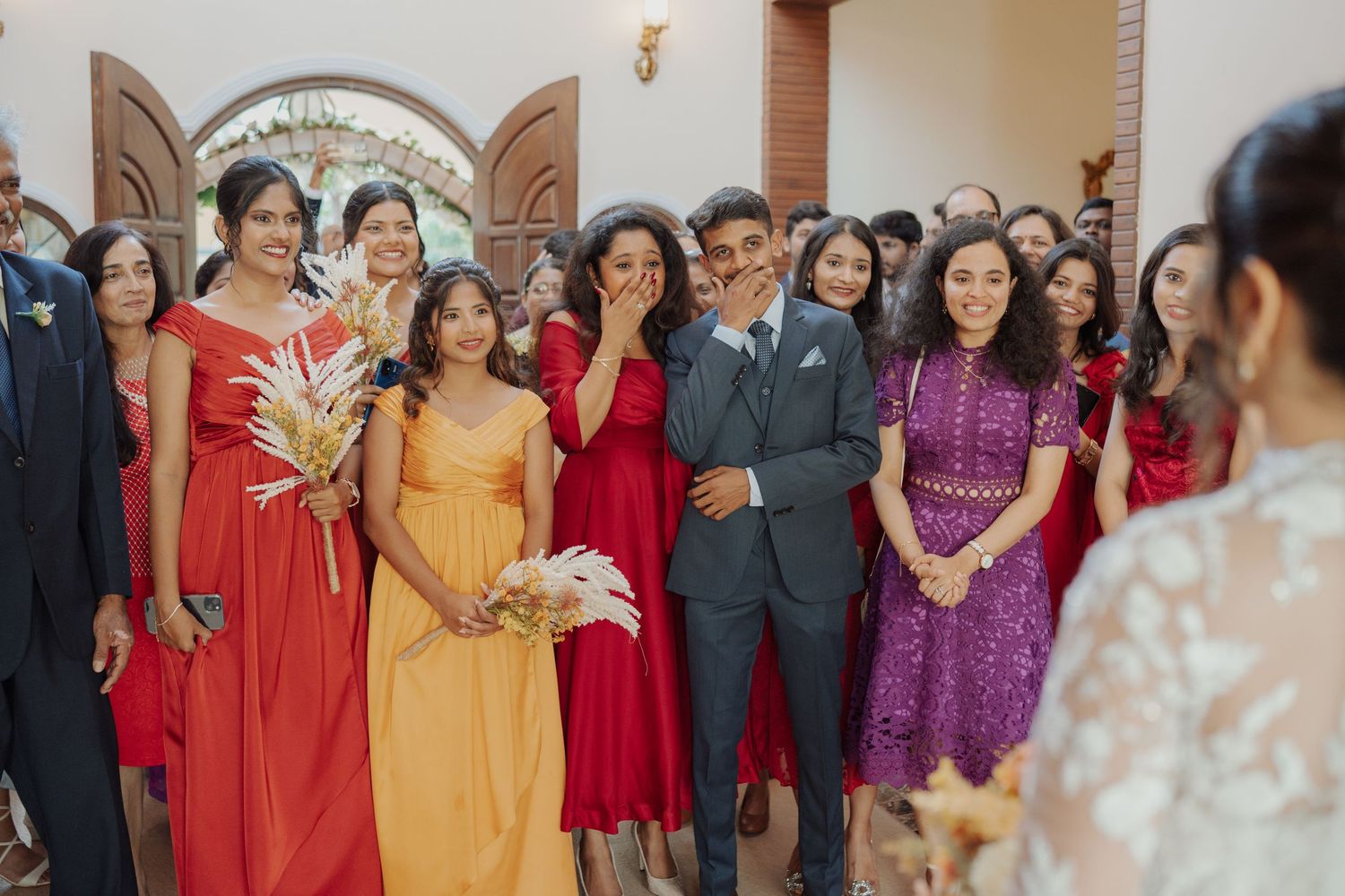 Wedding guests in colorful dresses and suits gather around a smiling groom, with one woman wiping happy tears during the celebration.