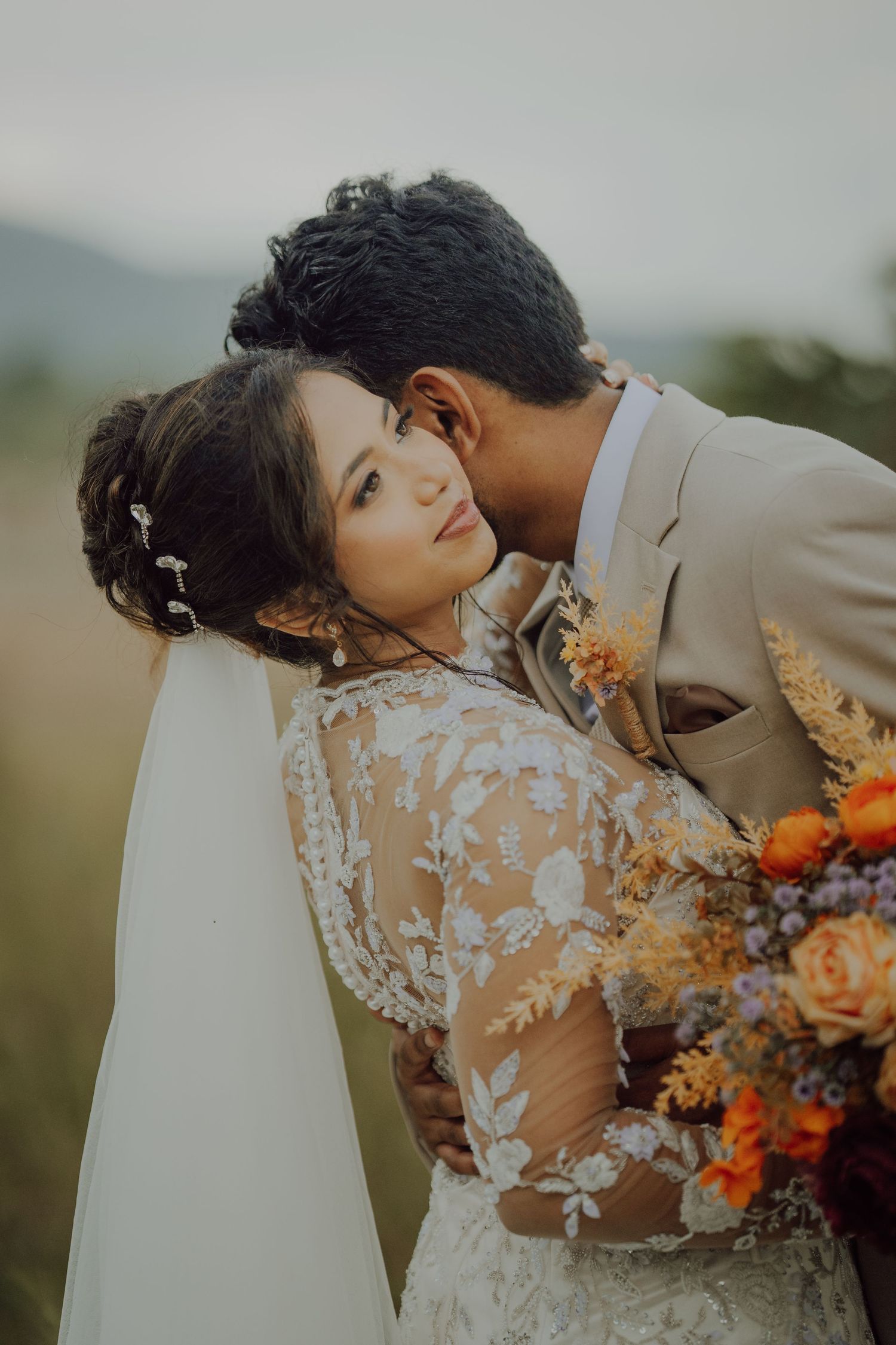 Bride in a lace wedding gown and veil embraces her groom in a tan suit, holding an orange and peach floral bouquet outdoors.