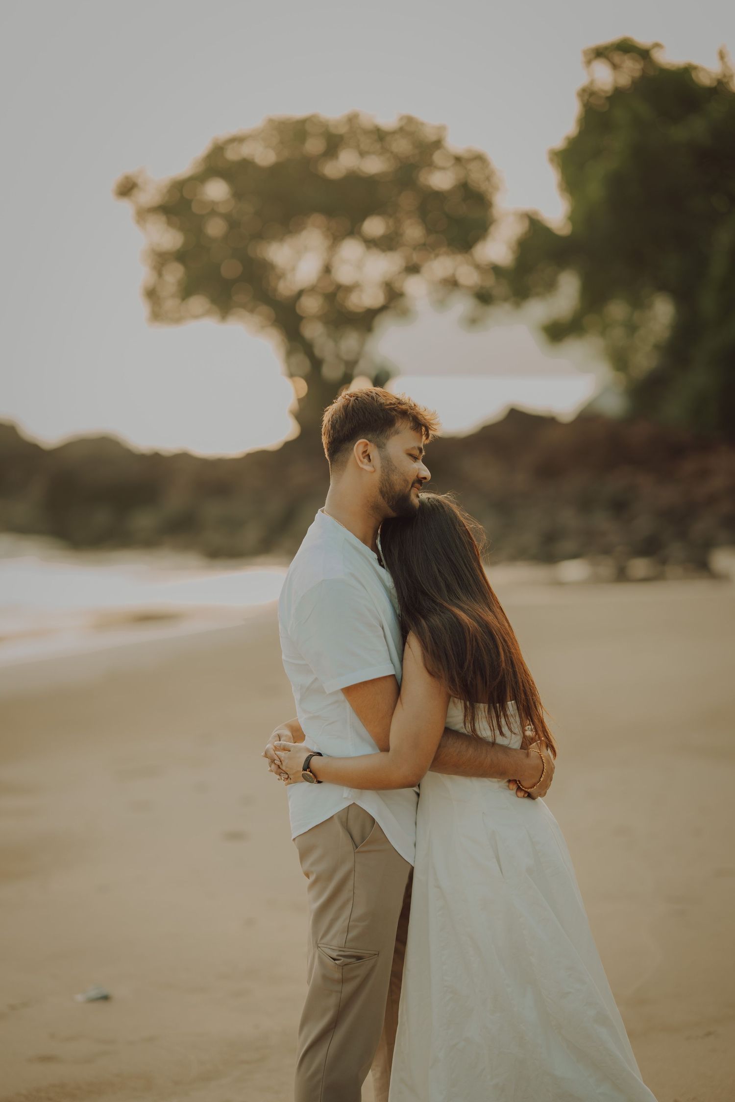 A couple embraces tenderly on a sandy beach during golden hour as sunlight filters through trees in the background.