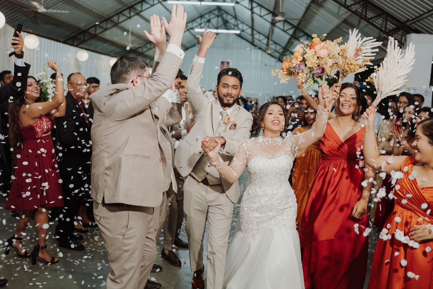A bride and groom celebrate with cheering wedding guests in a festive hall as confetti flies and an orange bouquet is raised.