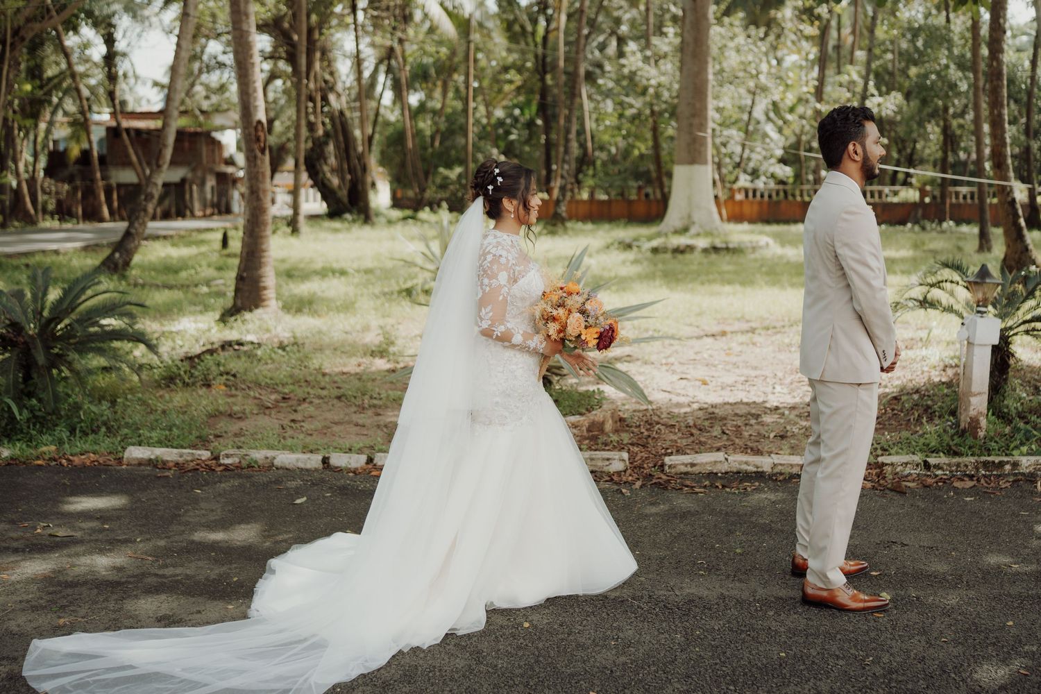 Bride in a long white lace gown holding a bouquet stands on a path as the groom in a beige suit waits among trees for a first look.