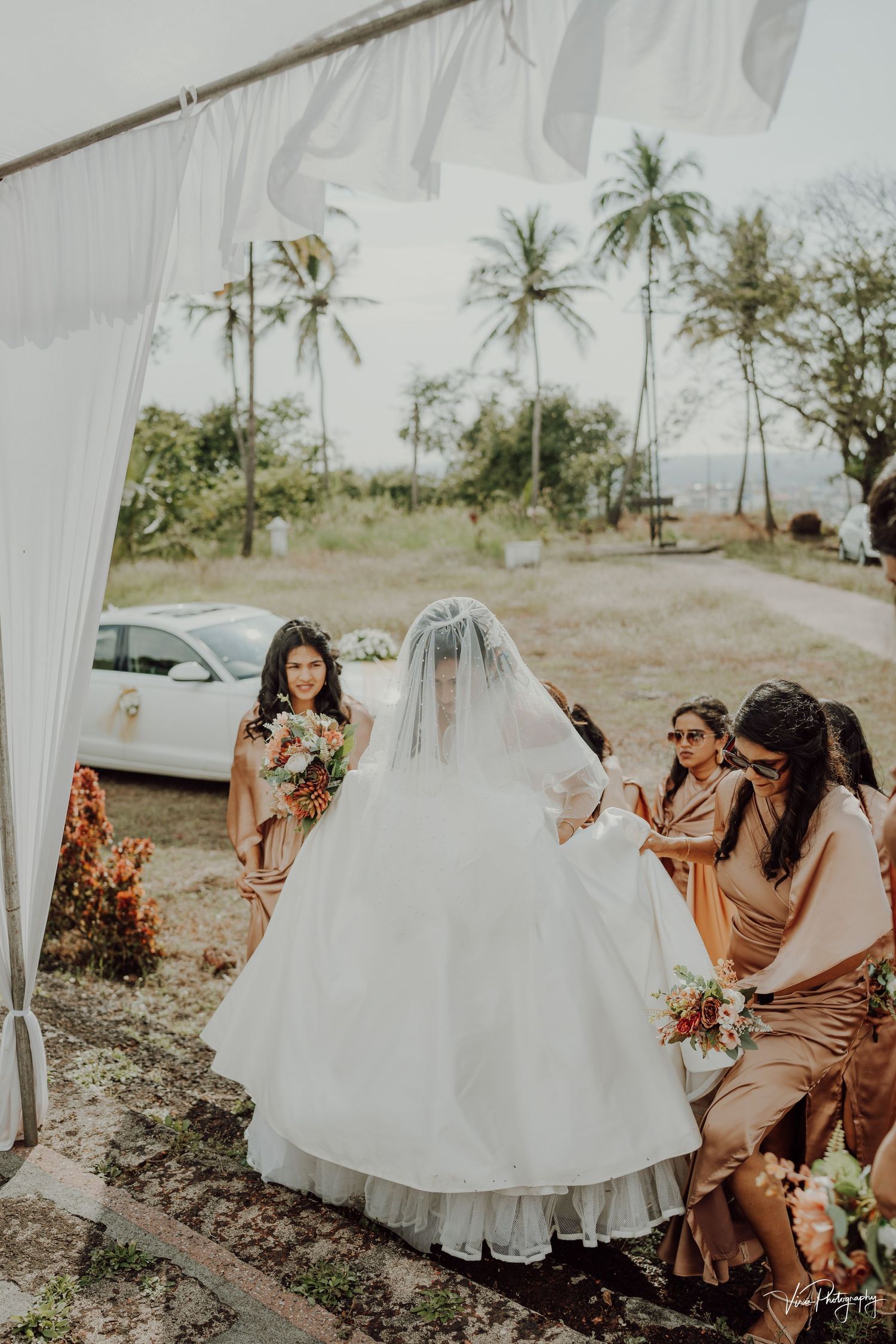 A bride in a white wedding dress and veil stands with several attendants at a tropical outdoor venue with palm trees.
