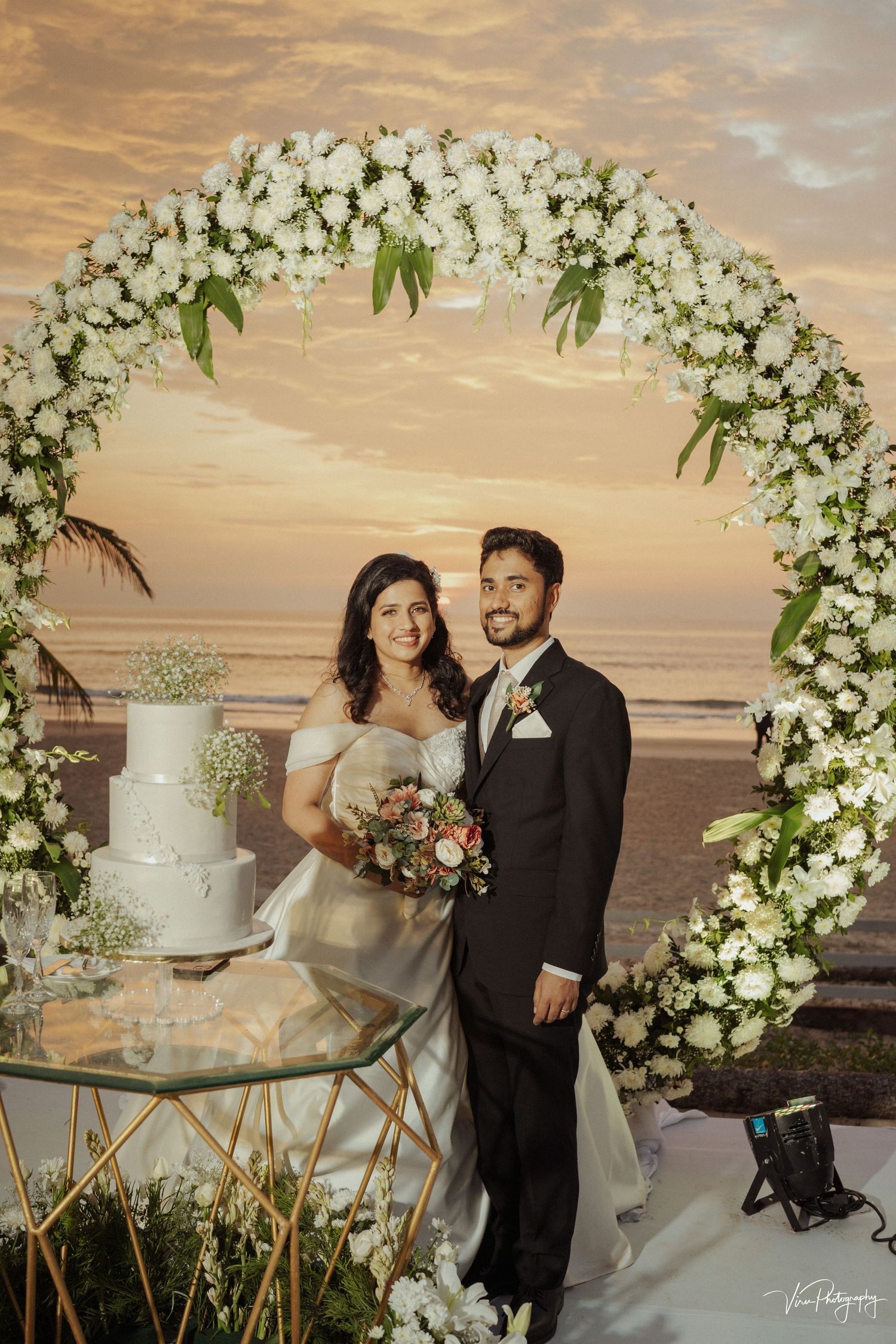A wedding ceremony setup on a beach at sunset with an elegant floral arch and white cake on a gold stand.