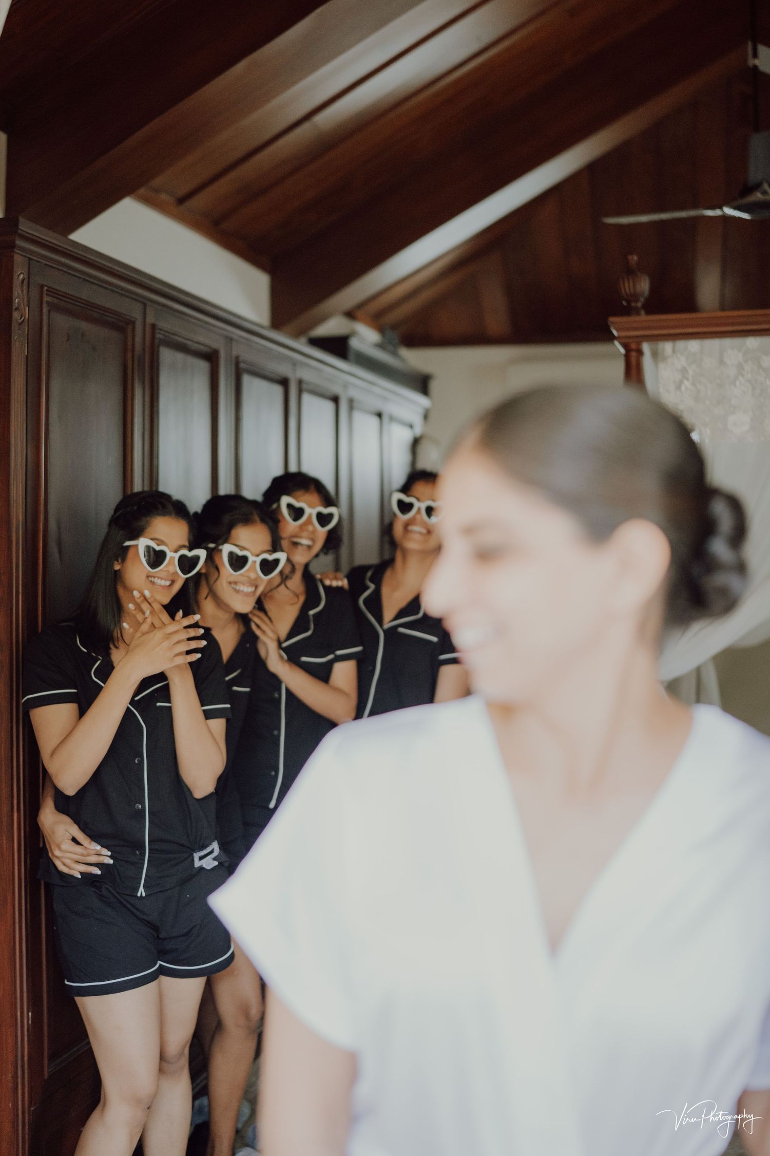 A group in matching black outfits and sunglasses pose playfully behind someone wearing white in an indoor setting.