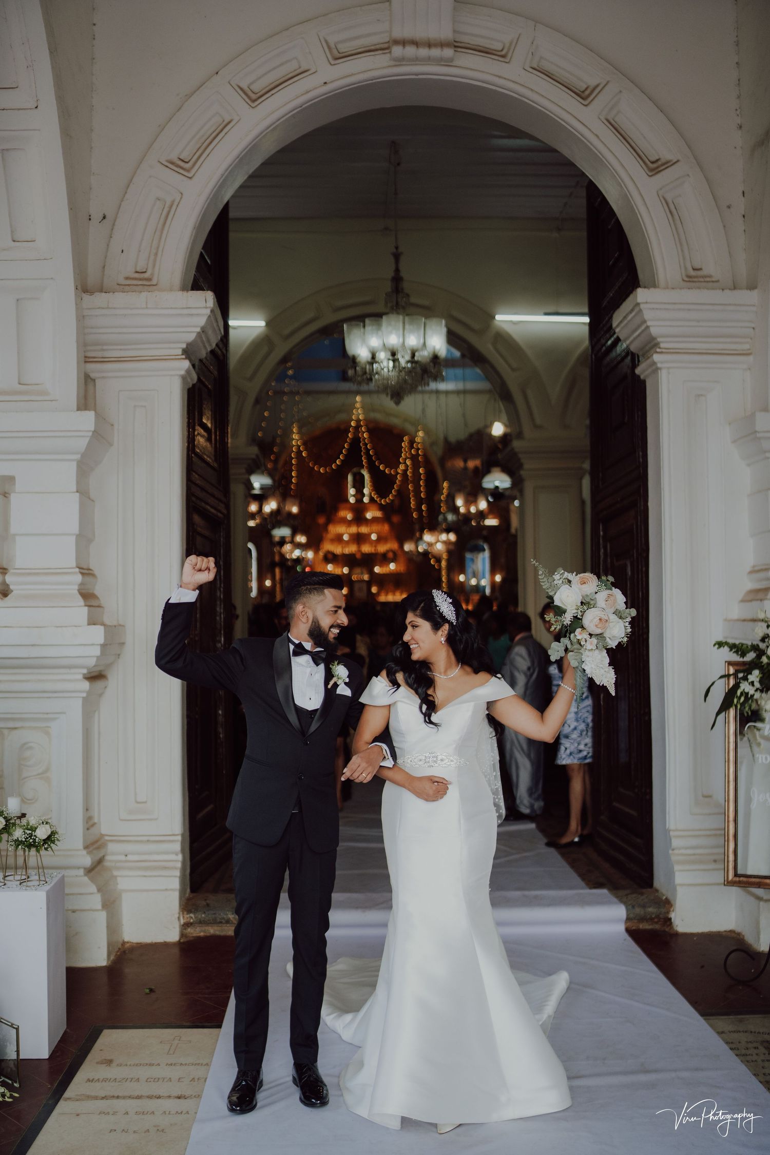 A white-arched church entrance with elegant chandeliers and holiday decorations frames a joyful wedding celebration.