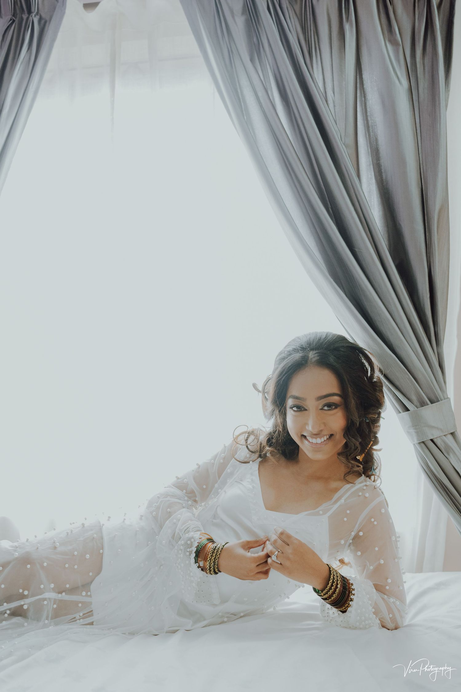 Ethereal bridal portrait in soft light with flowing white dress and curtains creating a dreamy atmosphere.