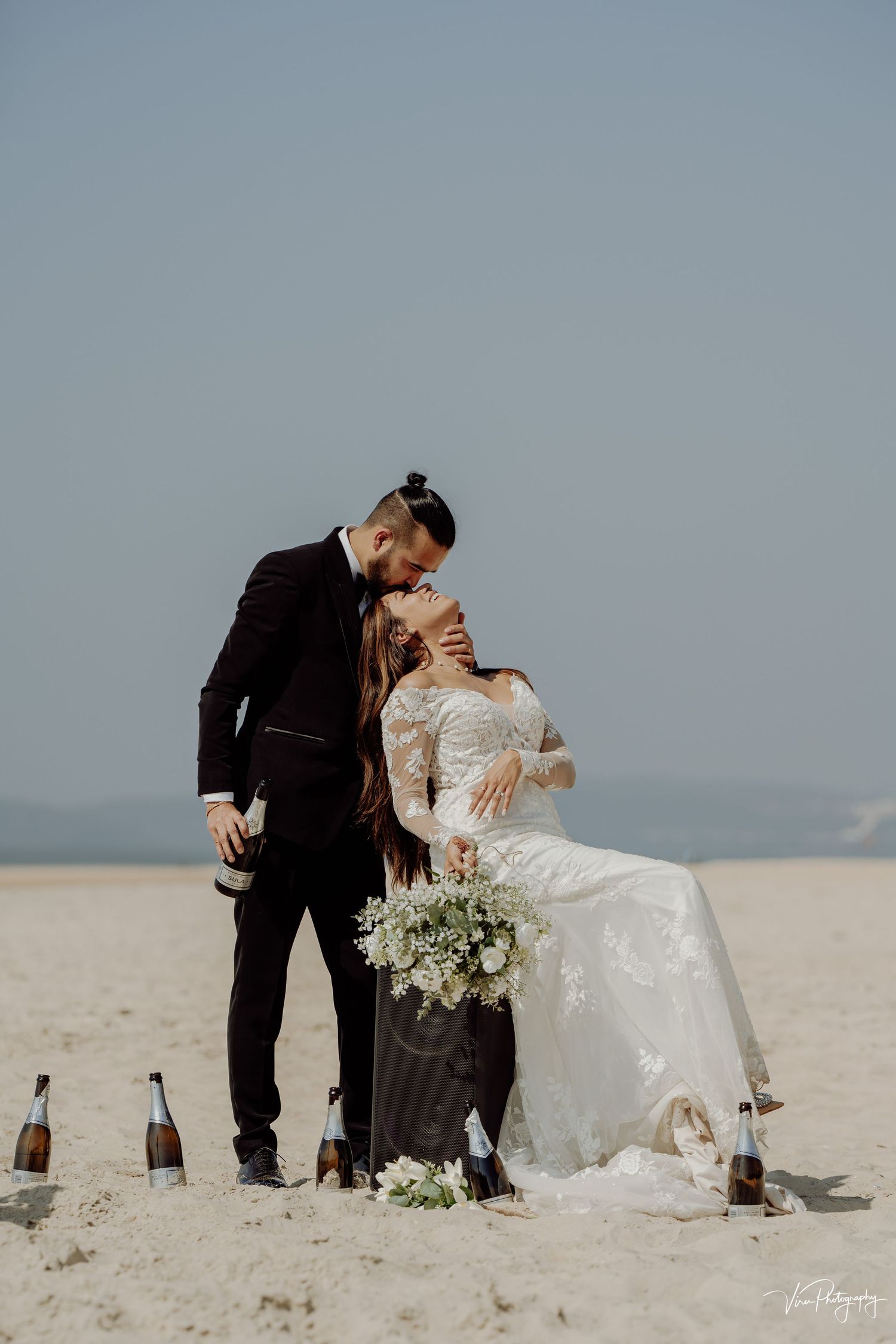 A romantic beach wedding scene with a couple embracing on the sand, surrounded by flowers and champagne bottles.