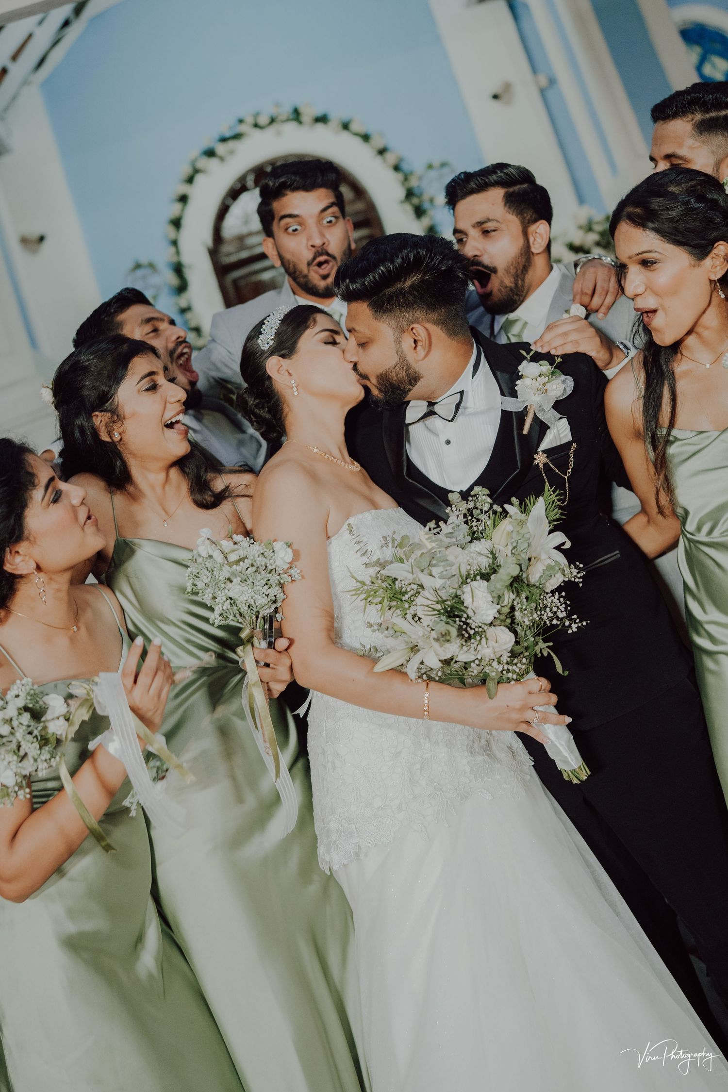 Wedding party in mint green bridesmaids dresses reacts joyfully as newlyweds share a romantic kiss.