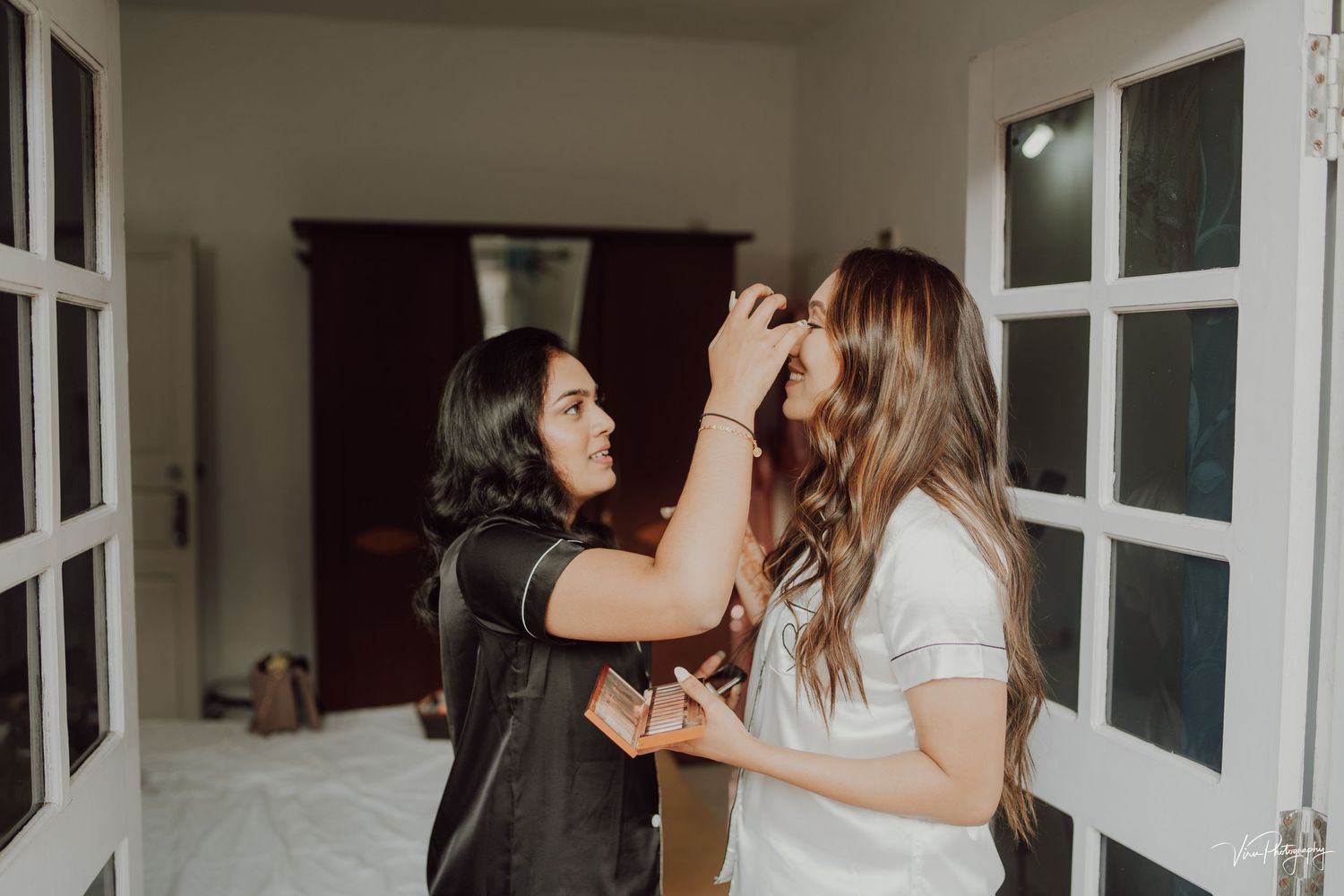 Two people having a fun moment as one applies makeup to the other in a bright interior room with white French doors.