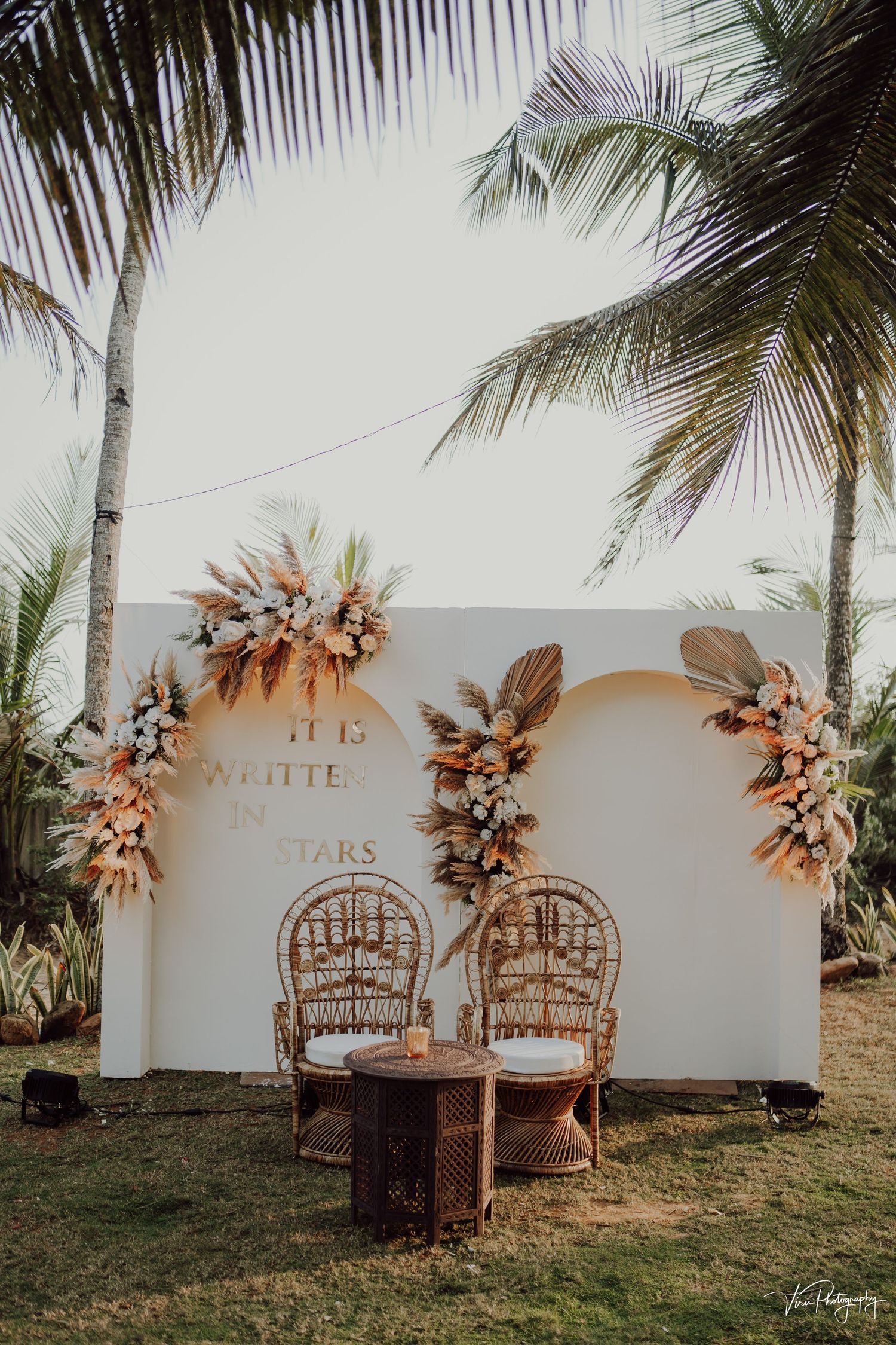 Rustic peacock chairs and floral arch setup for a tropical beach wedding with palm trees and white wall backdrop.