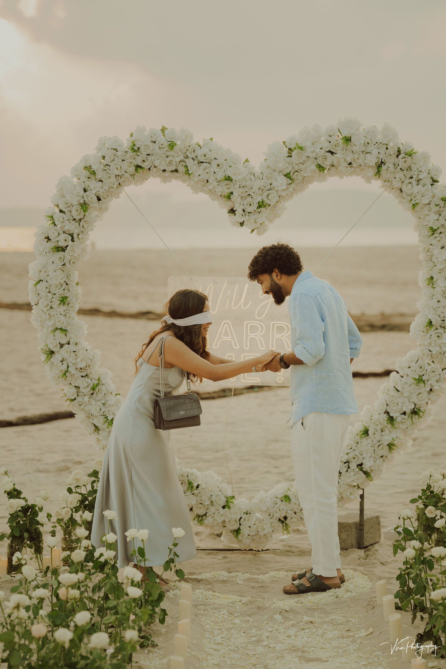 A couple stands beneath a heart-shaped floral arch on a beach at sunset, sharing an intimate moment together.