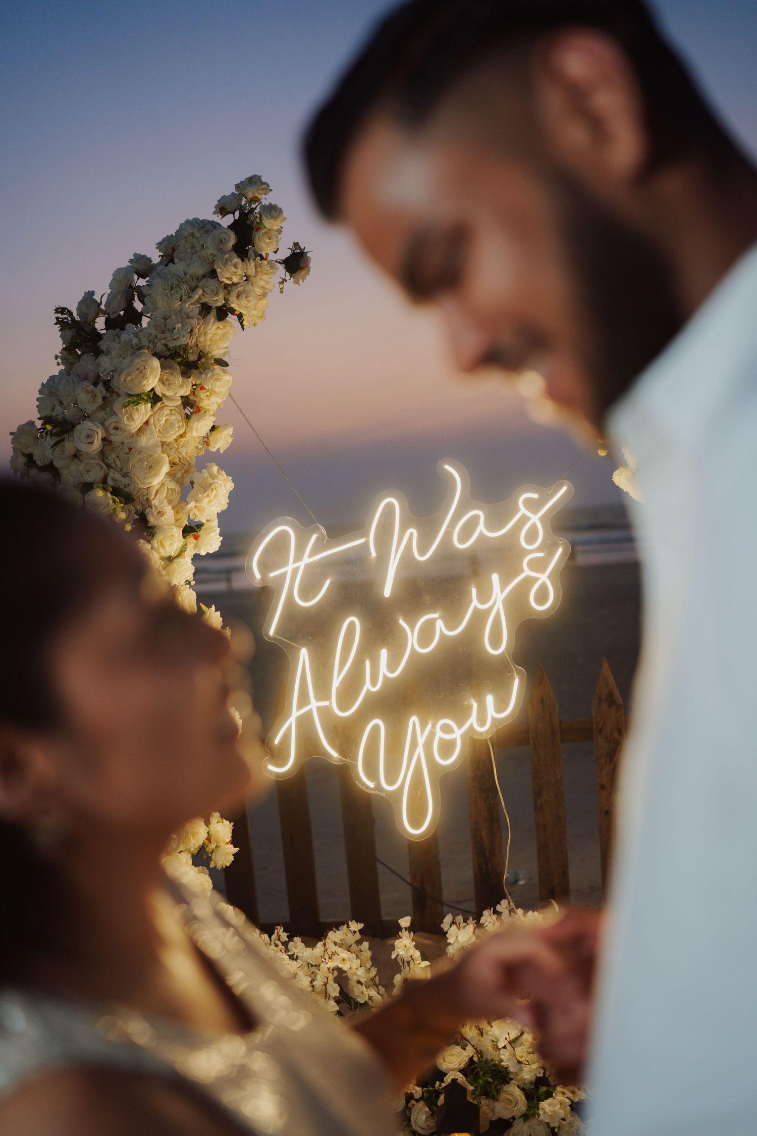 Romantic couple at sunset wedding with neon sign reading It Was Always You and white floral arch.