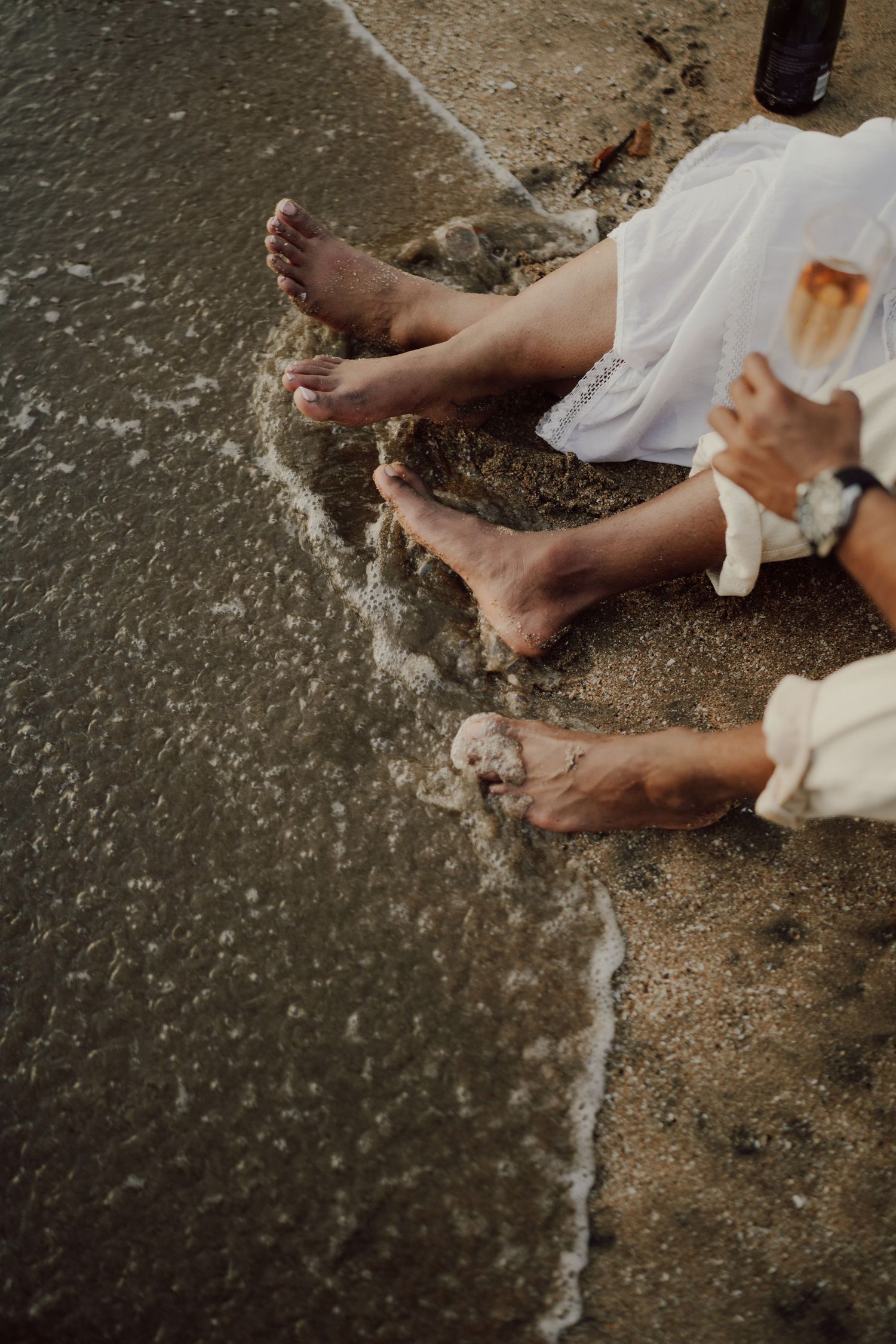 Two pairs of bare feet dangle over the edge of a surface while waves splash below, with someone holding a drink.