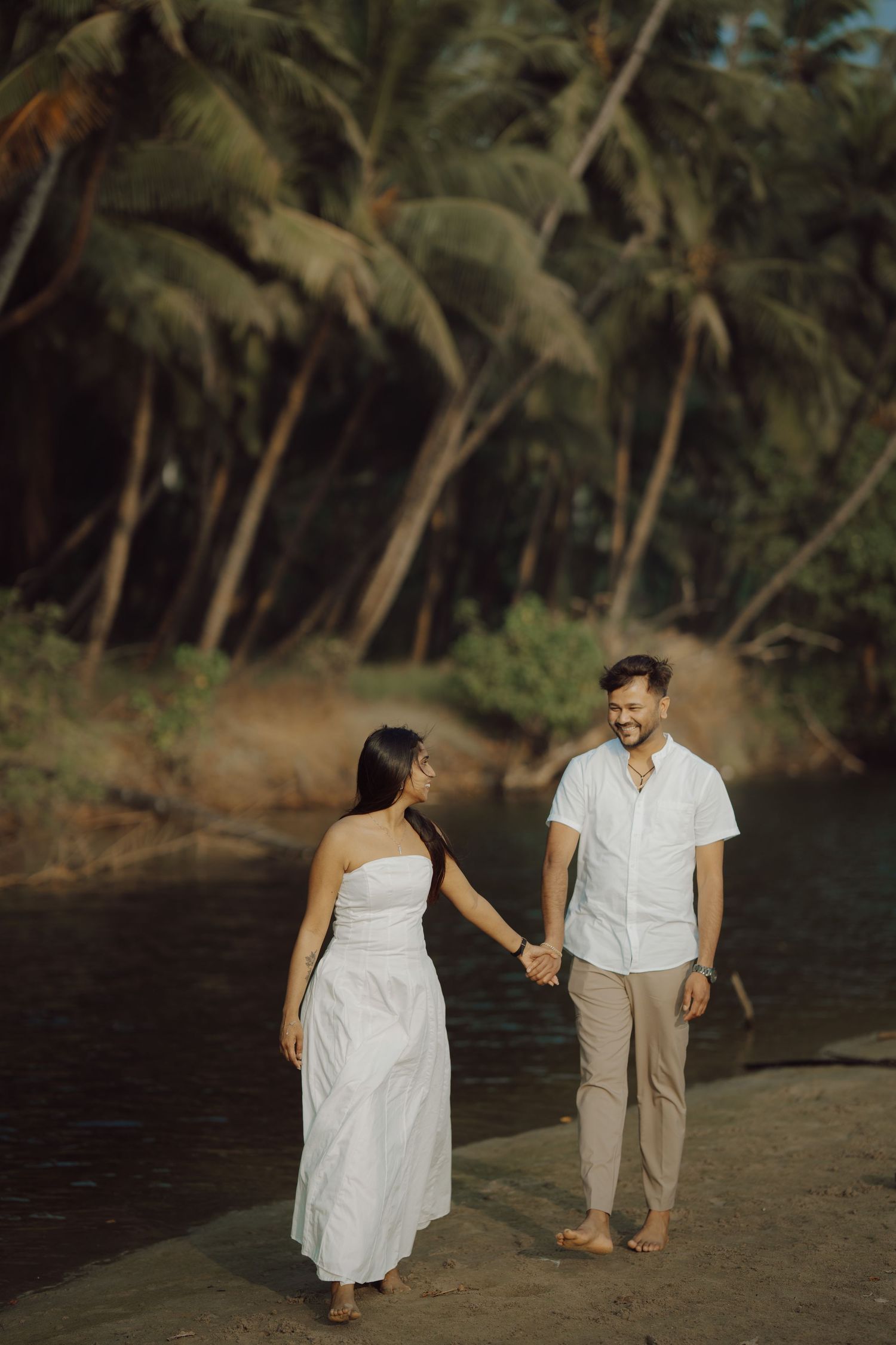 A couple in white attire walks along a tropical beach lined with palm trees at sunset.