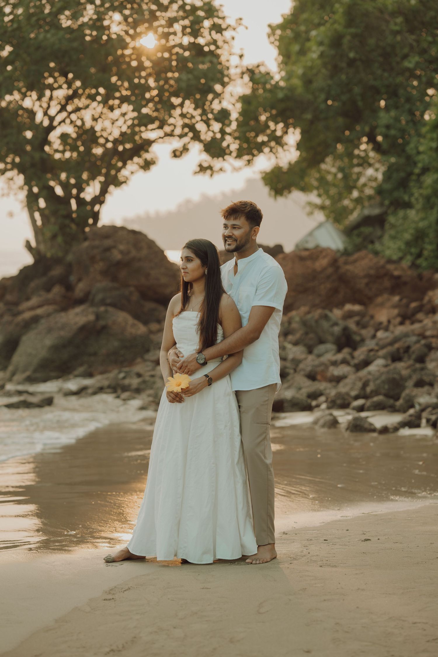 A couple embraces during golden hour on a beach with rocky cliffs and trees in the background.