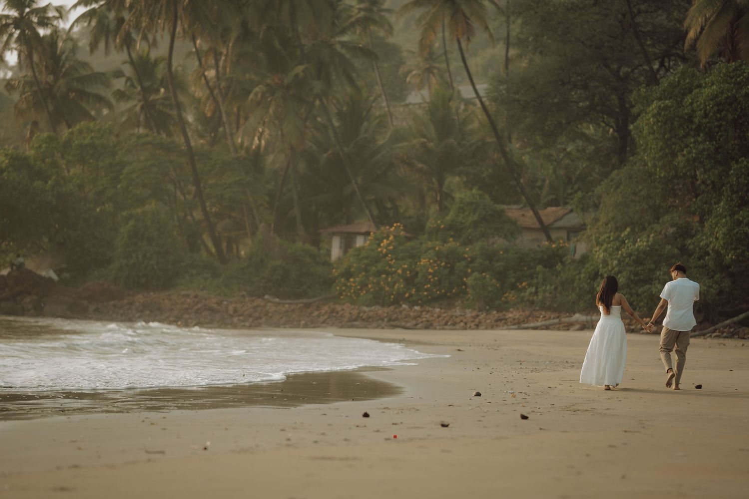 A couple walks along a tropical beach with palm trees and misty atmosphere at sunset.