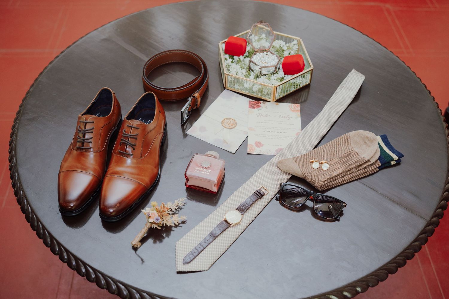 Groom's wedding day essentials including brown leather shoes, belt, tie, sunglasses, watch, socks and a floral boutonniere on a round table.