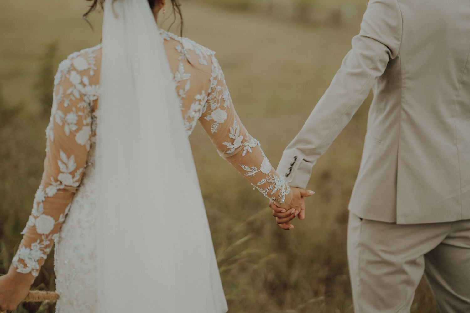 Bride in a lace-sleeved gown and veil holds hands with the groom in a beige suit while walking through a golden grass field.
