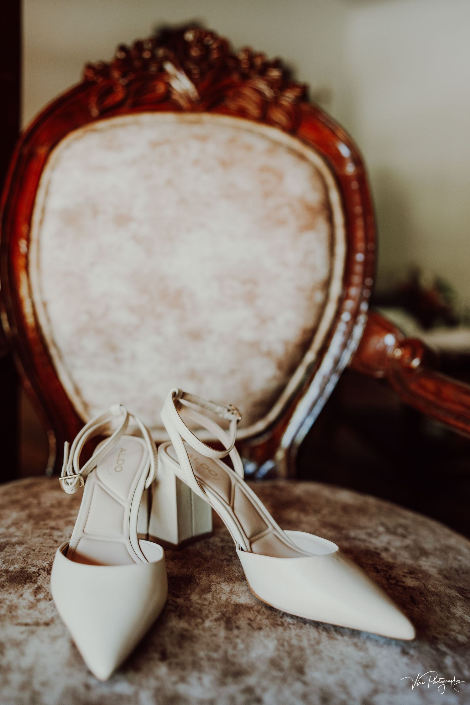 Elegant white bridal shoes with straps displayed against an ornate vintage chair with upholstered backing.