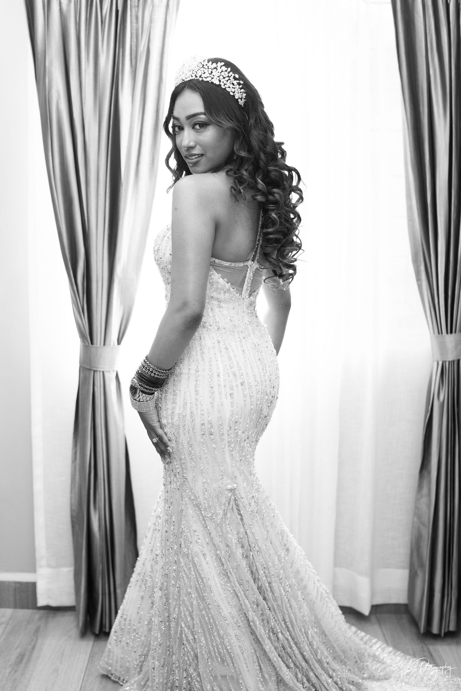 A black and white portrait of a bride in a stunning mermaid-style lace wedding gown with long curled hair and tiara.
