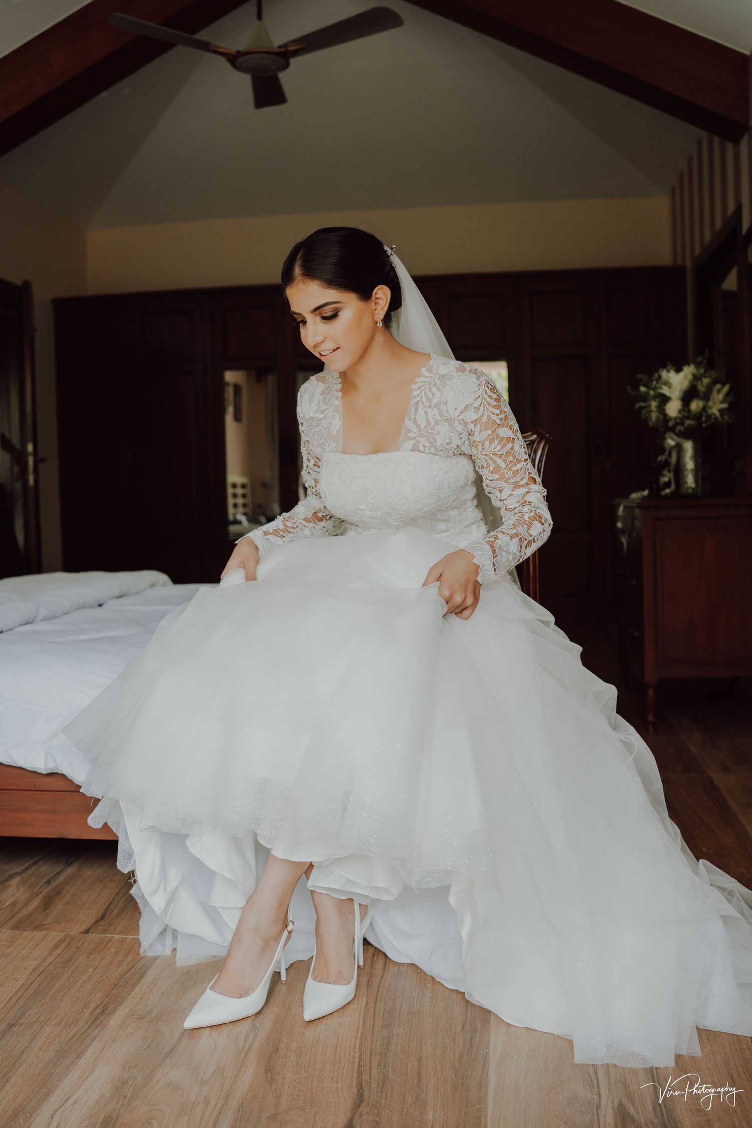 A person in an elegant white lace wedding gown with long sleeves adjusts their white heels in a rustic bedroom setting.