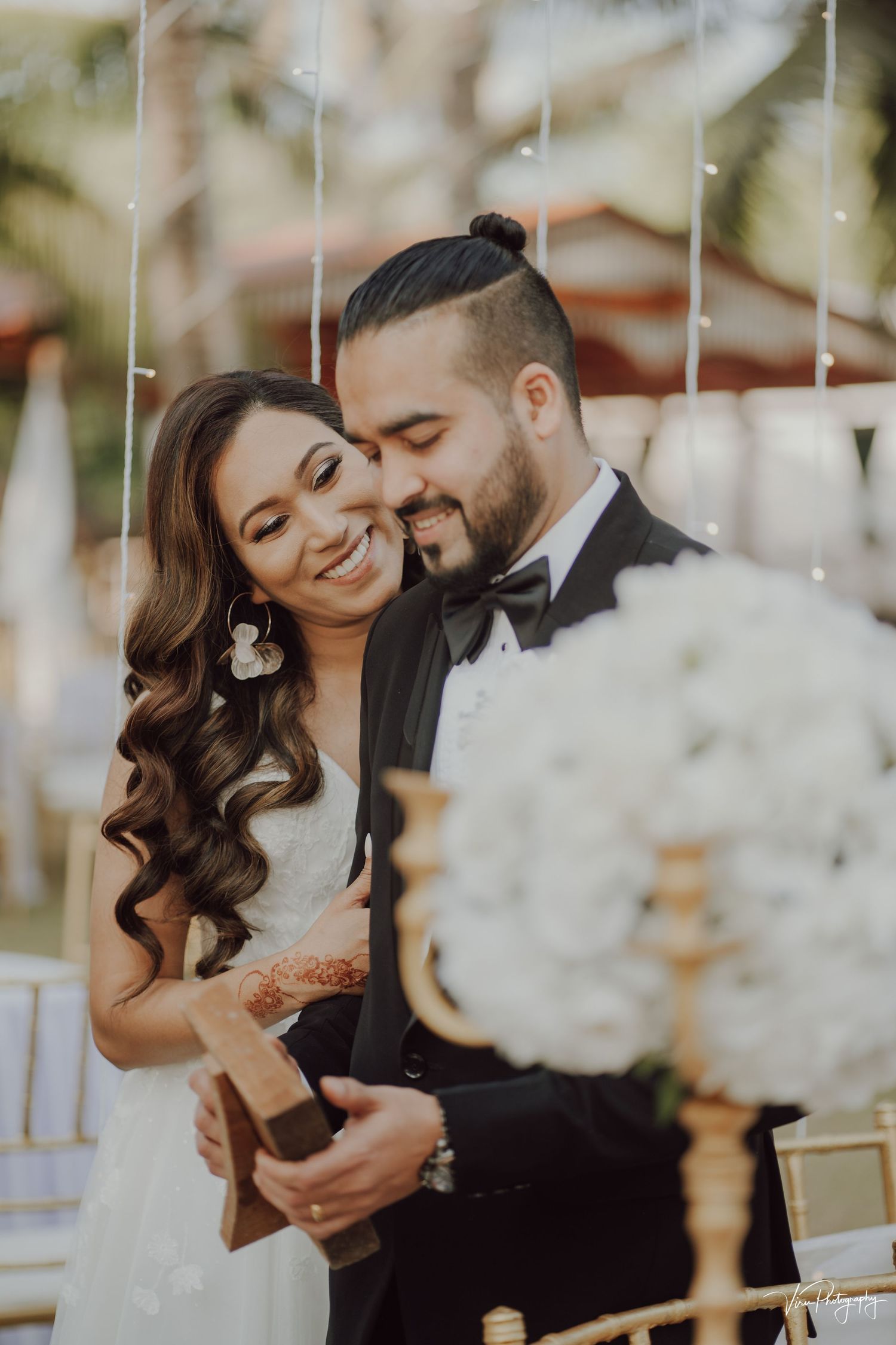 A romantic wedding moment captured as a couple shares a tender embrace amidst decorative white flowers and string lights.