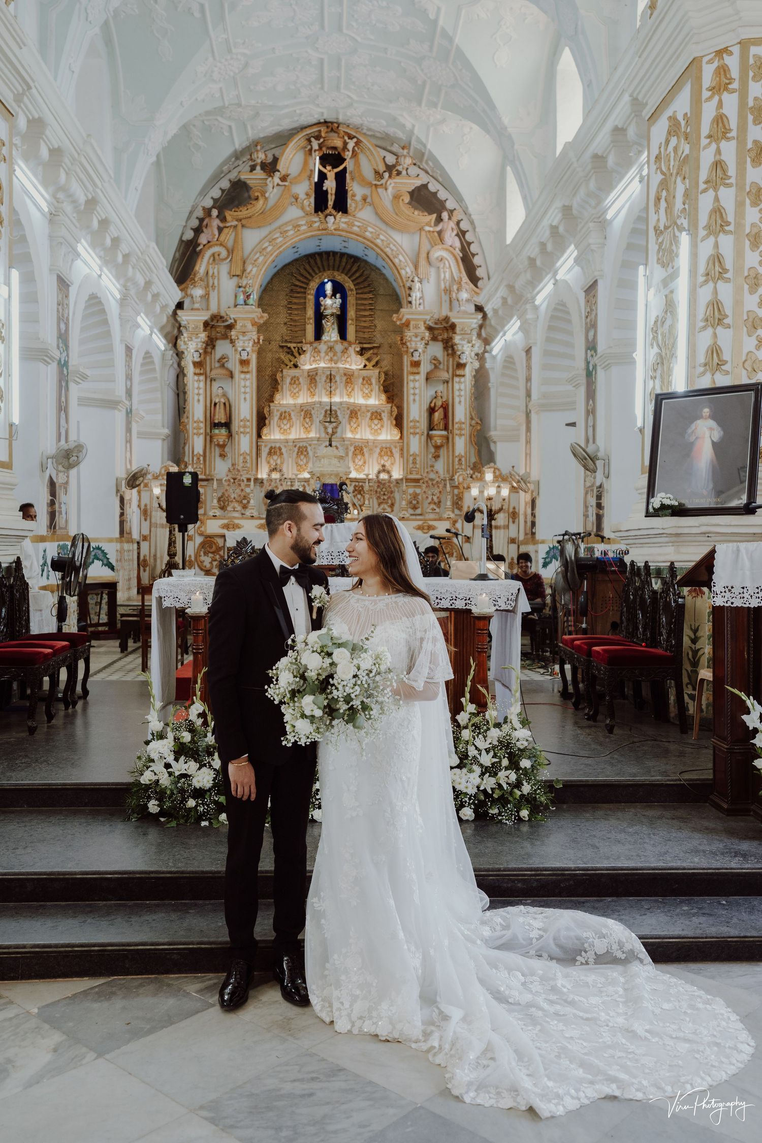 A romantic wedding ceremony inside an ornate white cathedral with baroque altar and floral arrangements on the steps.