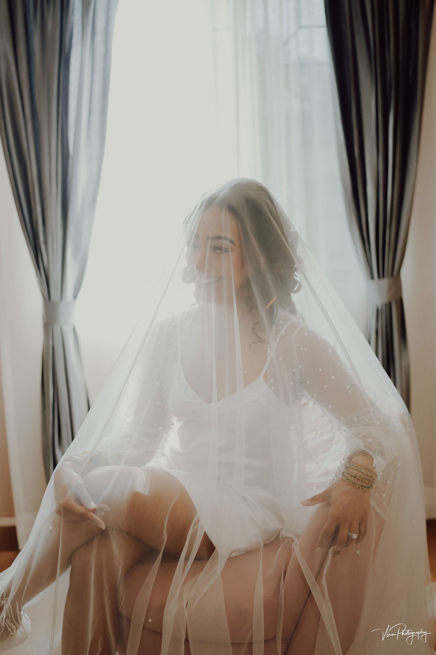An ethereal bridal boudoir photo with soft natural lighting through sheer curtains and a delicate white veil.