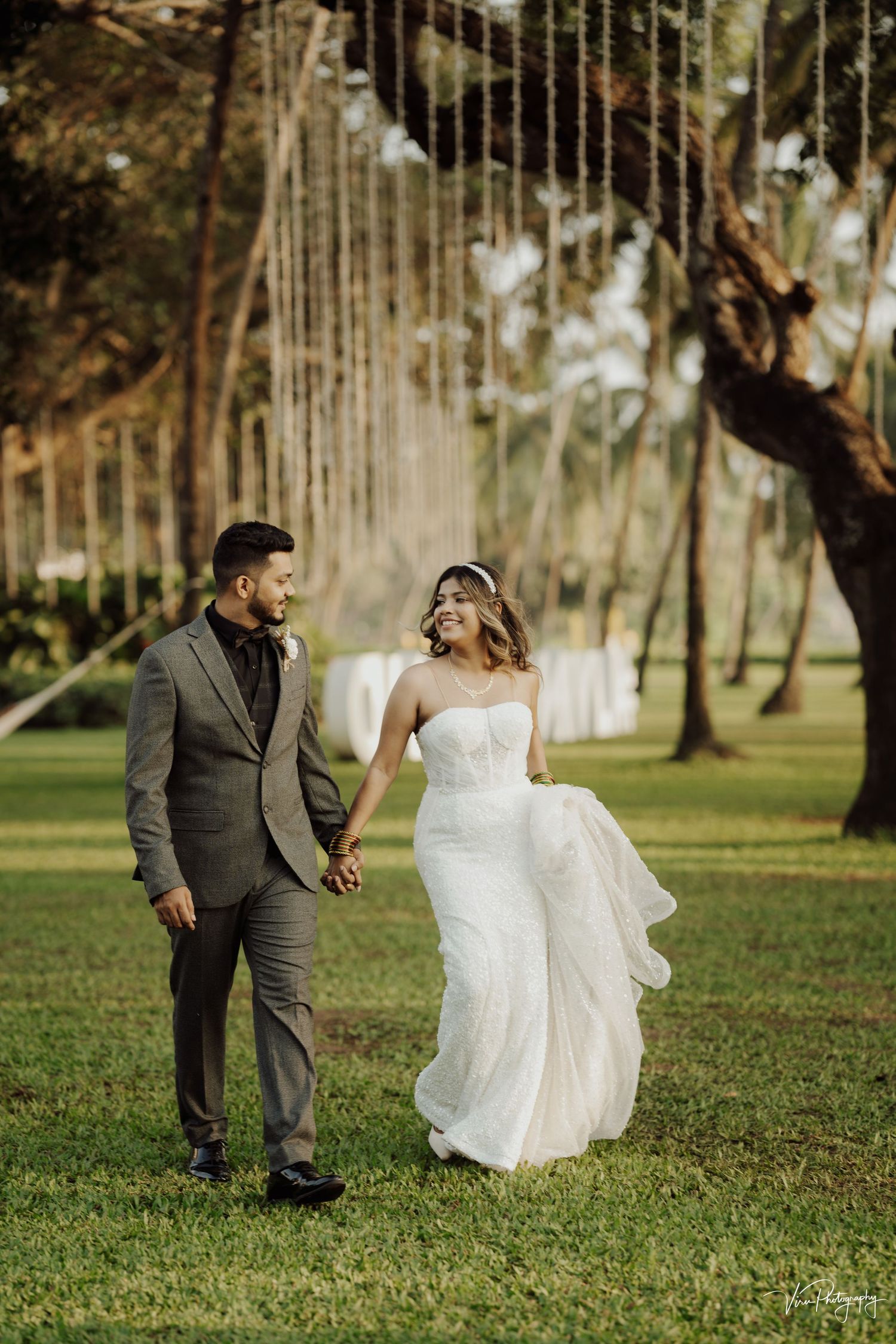 A couple in wedding attire walks hand in hand through a lush garden with palm trees and sunlit streaming through.