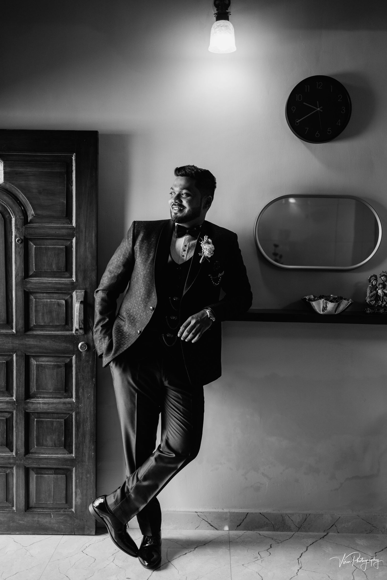Black and white photo of a well-dressed person leaning against a wall under a single light bulb with a clock and mirror.