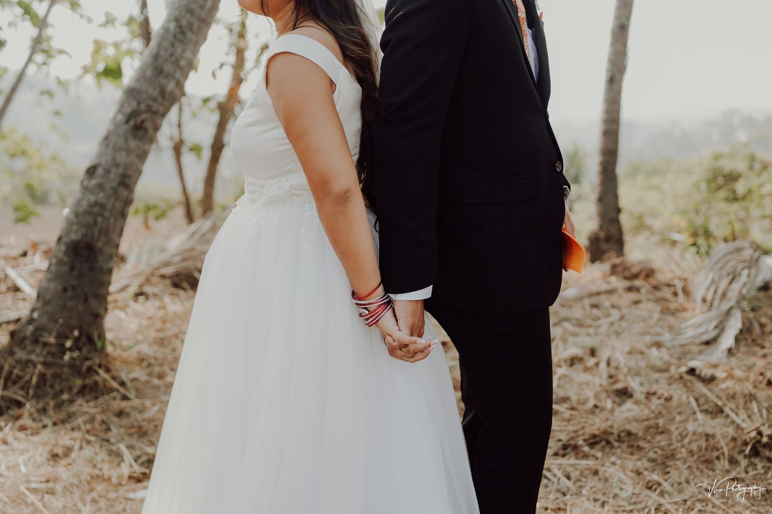 A bride in white gown and groom in black suit hold hands while standing together in a rustic outdoor setting.